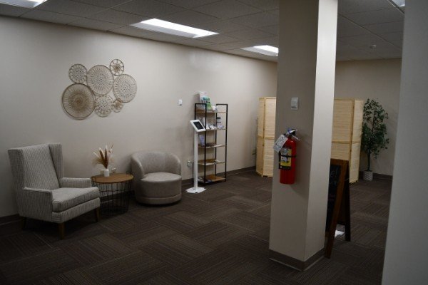 A waiting room with two armchairs and a round ottoman, a wall decoration of woven circular art, and a small shelf with informational materials. There is a fire extinguisher attached to a wall and partially visible wooden partitions in the background.