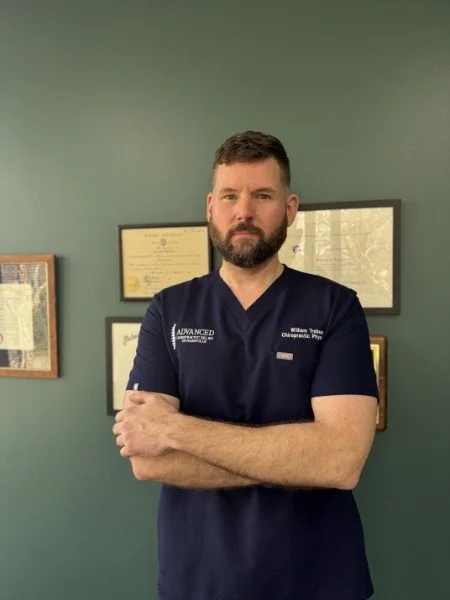 Man with short dark hair and beard standing with arms crossed in a medical office, wearing navy blue scrubs, with framed certificates on the wall behind him.