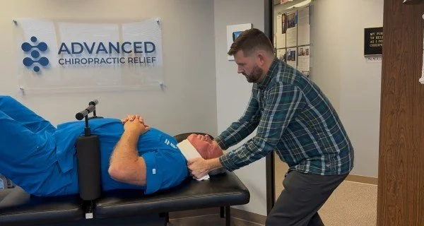 A chiropractor adjusting a patient lying on a chiropractic table in a clinic with a banner that reads ‘Advanced Chiropractic Relief,’ and a staff member standing nearby.