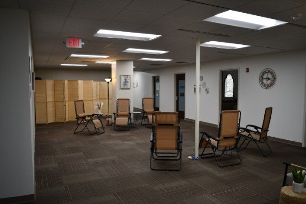 A spacious waiting room with several brown and black chairs arranged around the room. There is a small table with a potted plant, a wooden room divider, and a clock on the wall. The ceiling has fluorescent lighting and there are white walls with some artwork.