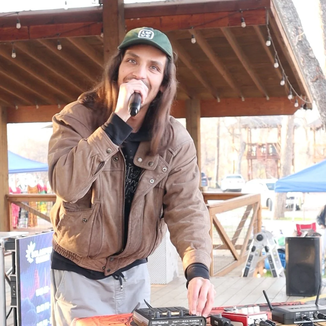 A young man with long brown hair, wearing a green cap and a brown jacket, speaking into a microphone while operating DJ equipment outdoors.