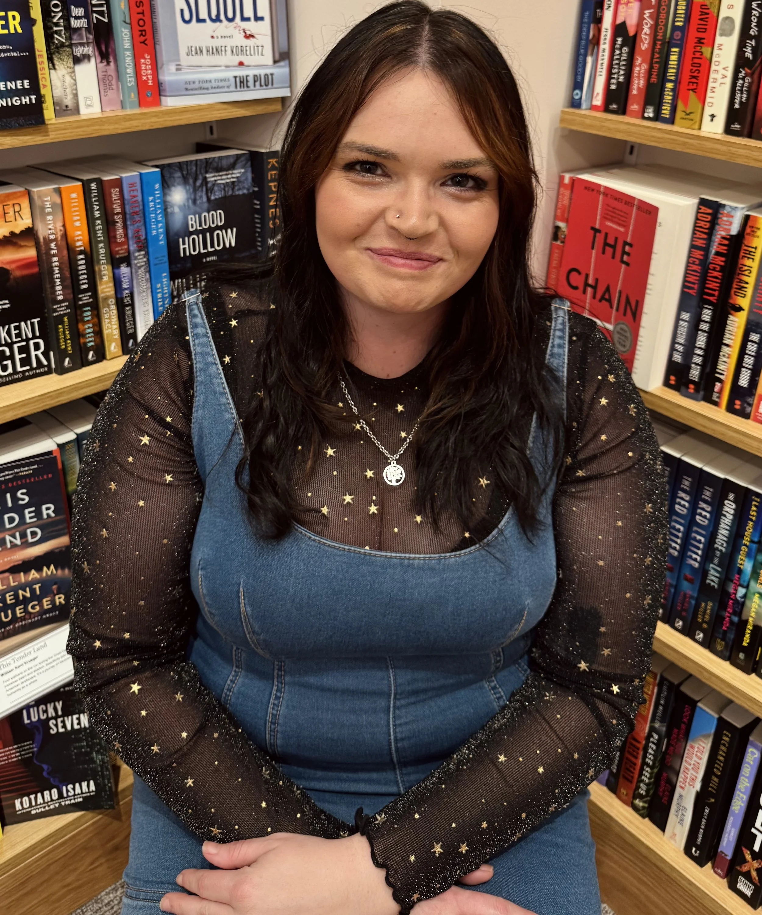 A young woman with long dark hair and a nose piercing sitting in a bookstore, surrounded by shelves of books.