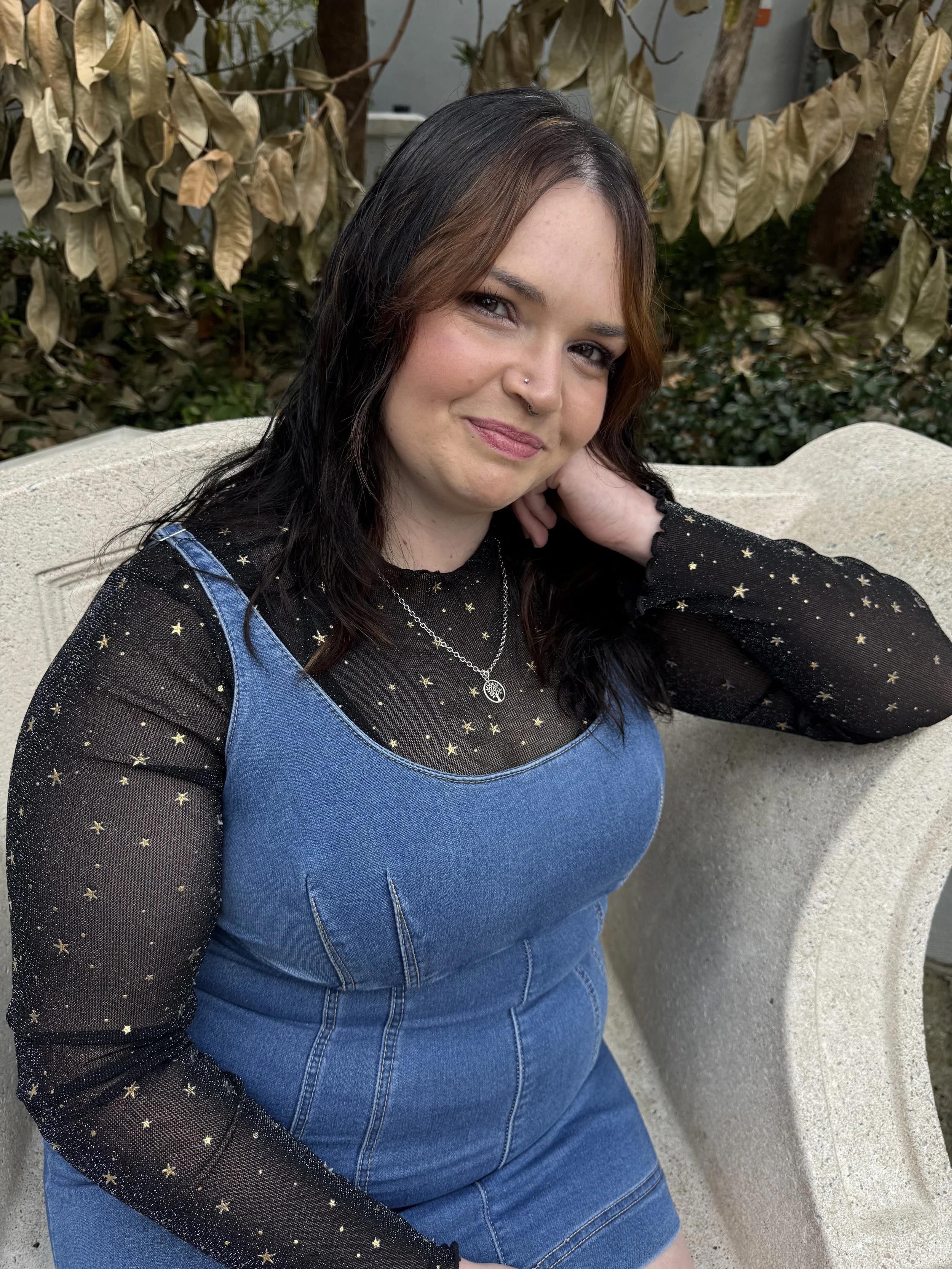 A woman with dark hair and light skin is sitting on a stone bench outdoors, wearing a black long-sleeve top with gold stars and a blue denim dress, surrounded by brown and green foliage.