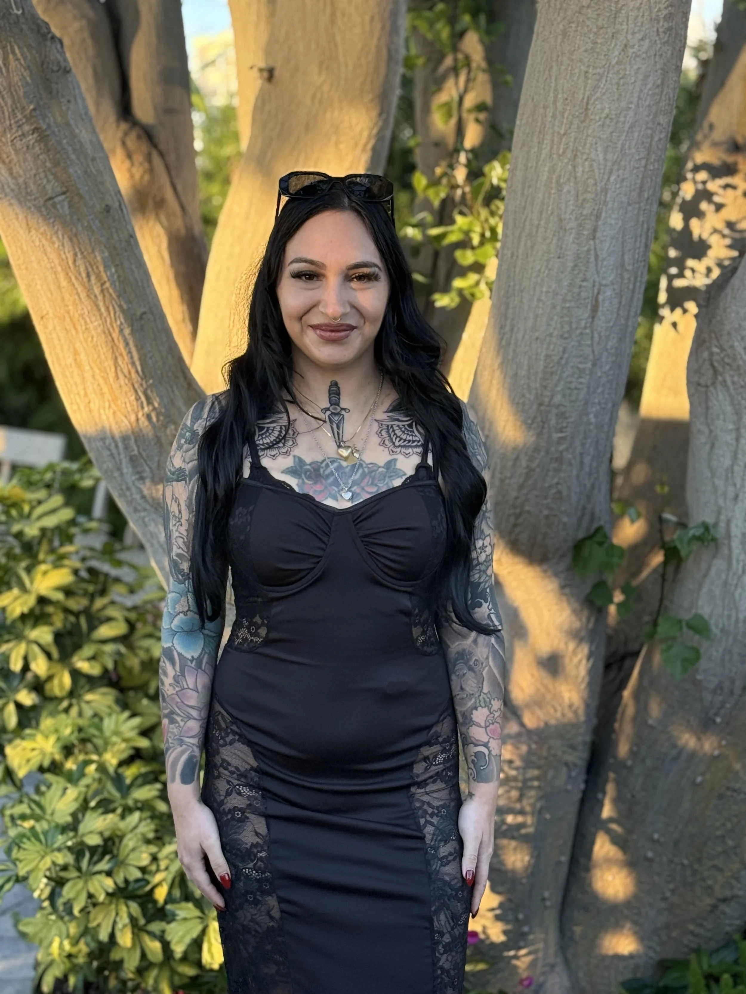 A woman with long black hair, tattoos on her arms and chest, wearing a black lace dress, and sunglasses on her head, standing in front of a large tree outdoors