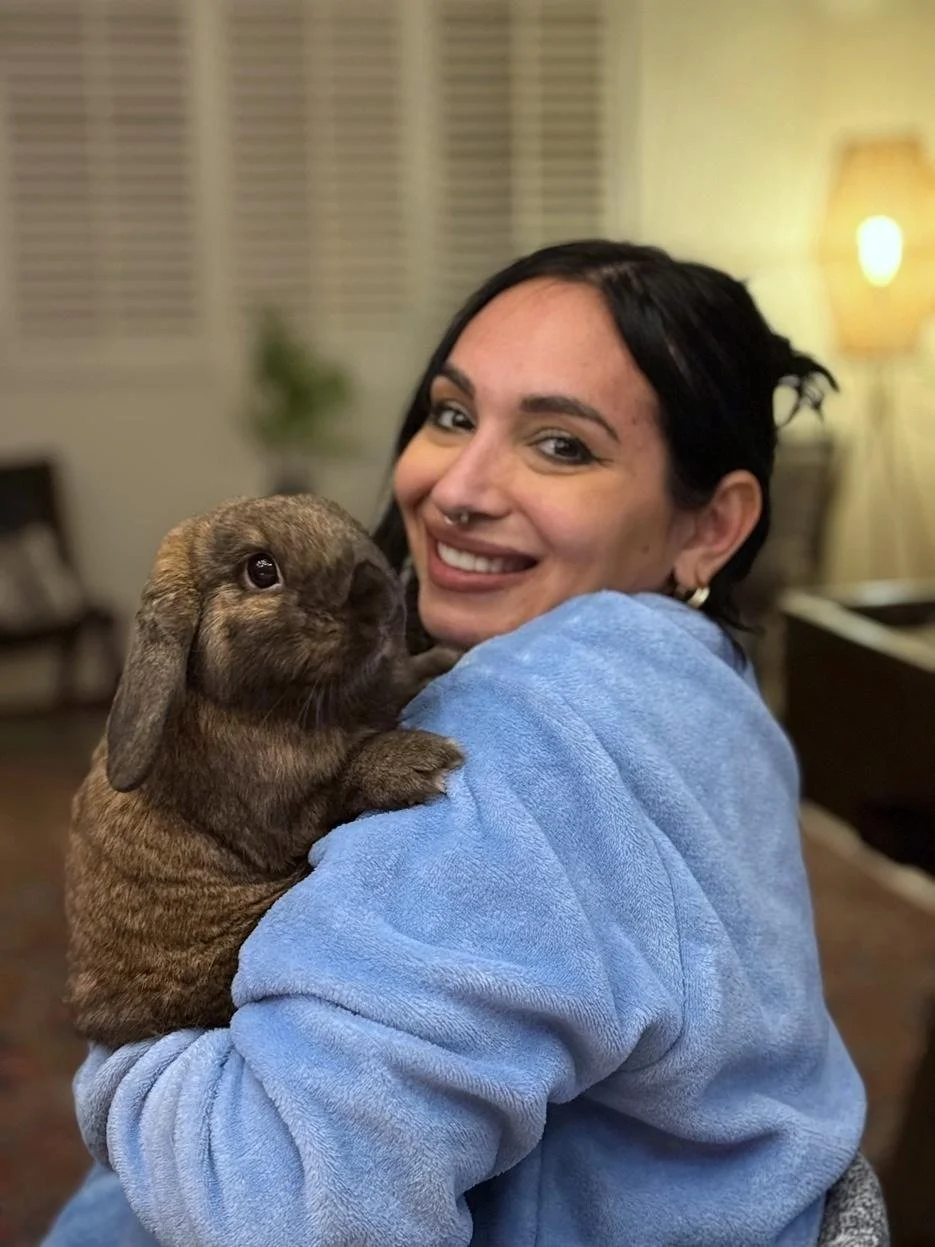 A woman with black hair, a septum piercing, and earrings, smiling and holding a brown baby rabbit indoors.