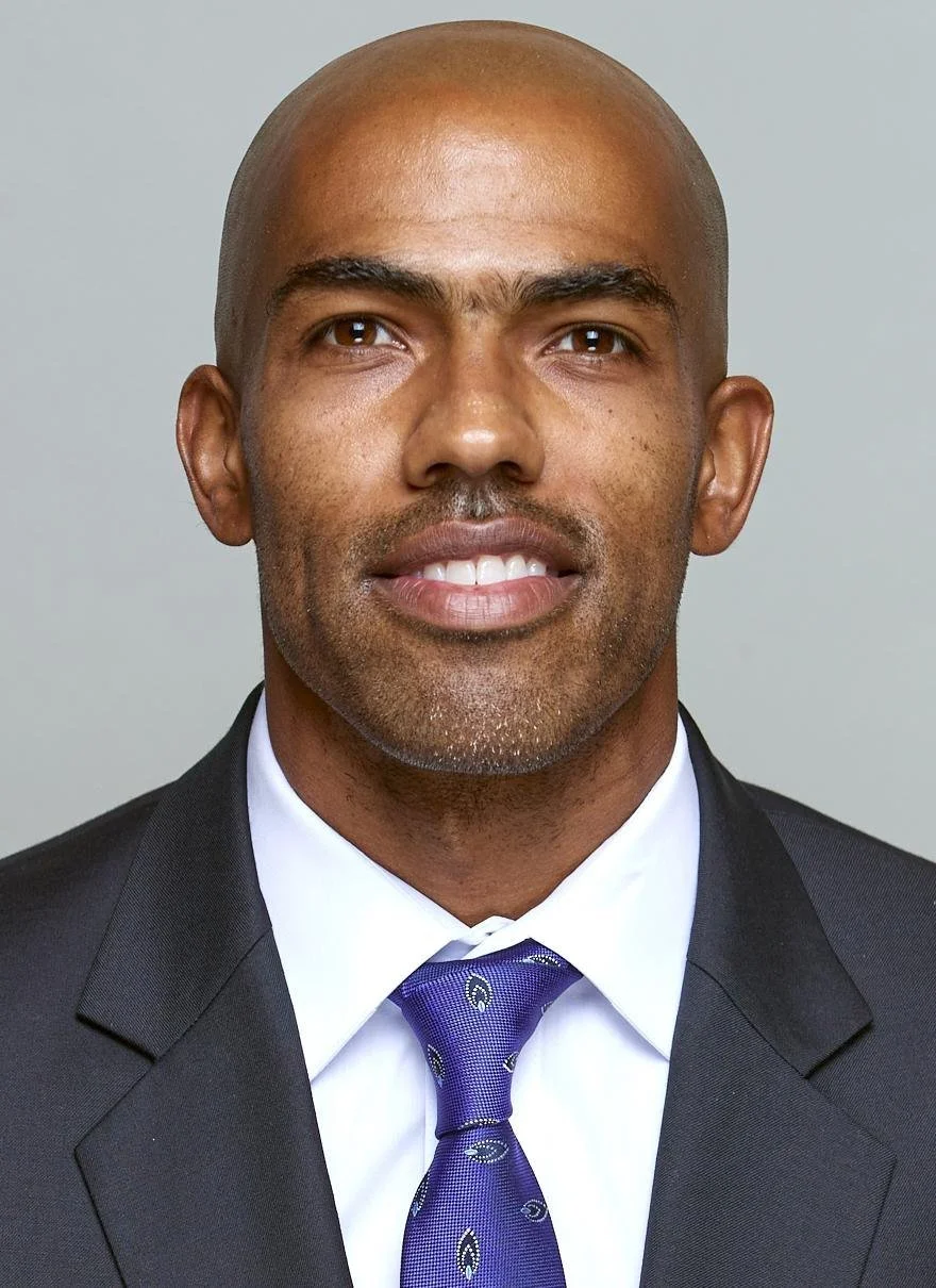 Close-up of a bald man in a suit smiling, wearing a white shirt and purple tie, against a plain light background.