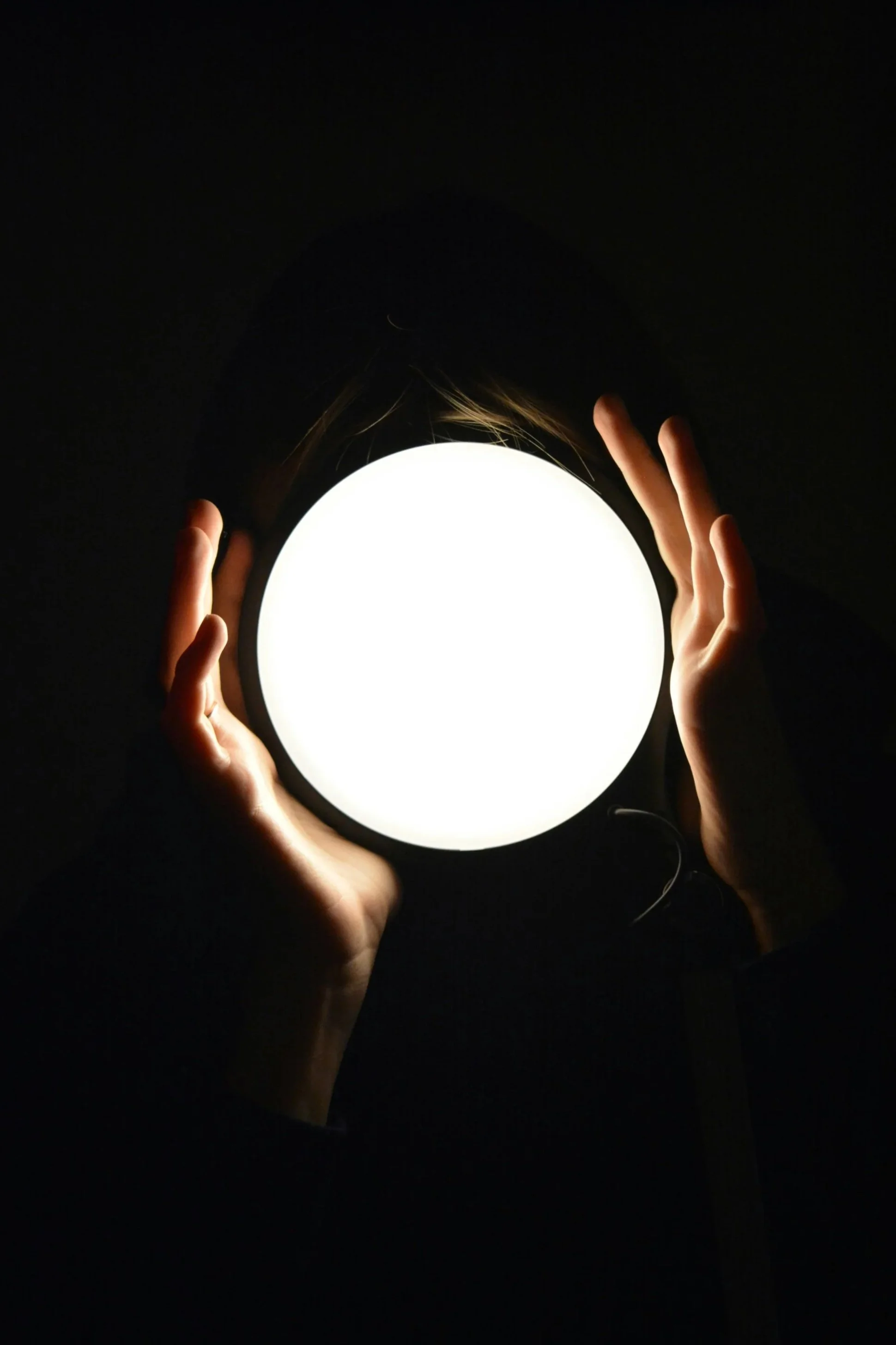 Person holding a bright, white, circular light source in darkness with hands framing the light.
