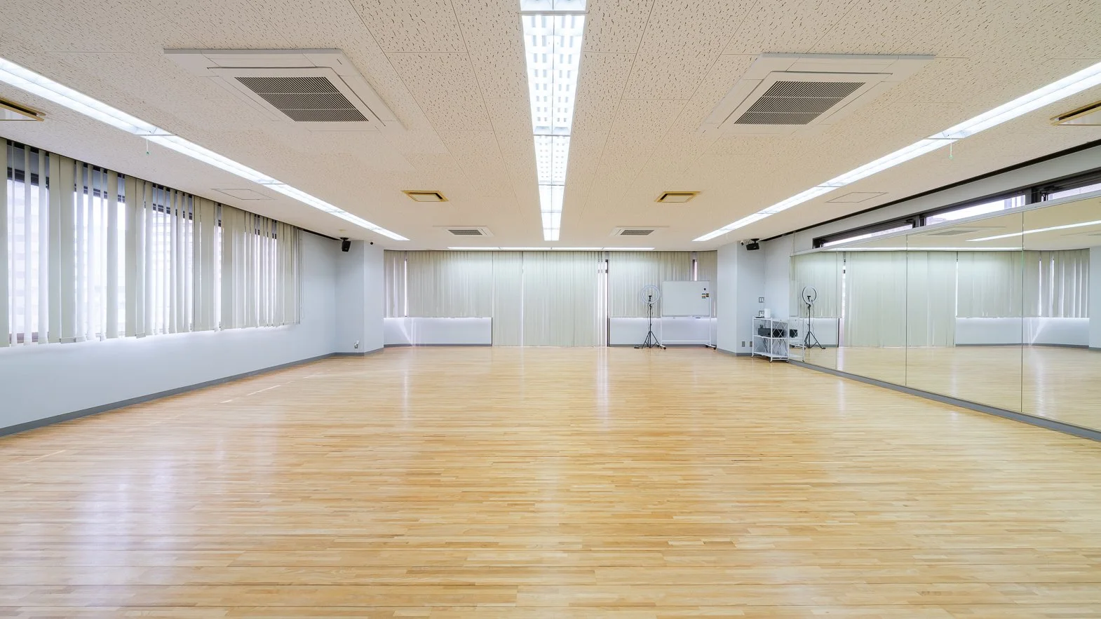 Empty dance or fitness studio with a light wooden floor, mirrored wall on the right, and large windows with vertical blinds on the left, bright ceiling lights, and minimal equipment in the back corner.