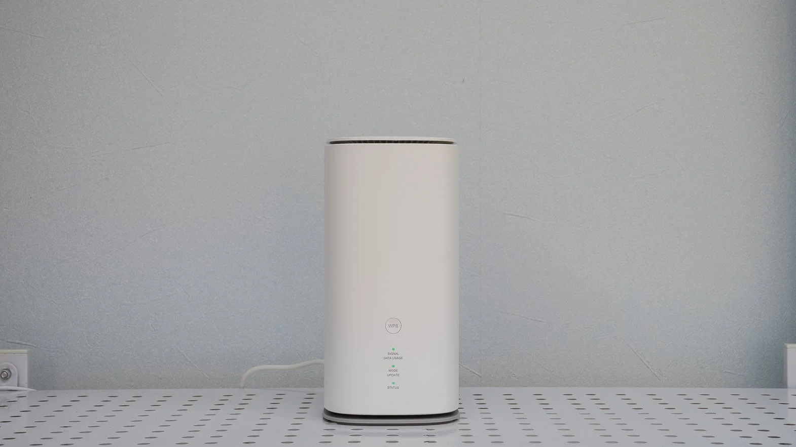 A tall white cylindrical electronic device with indicator lights and labels, positioned on a perforated white surface against a light gray textured background.