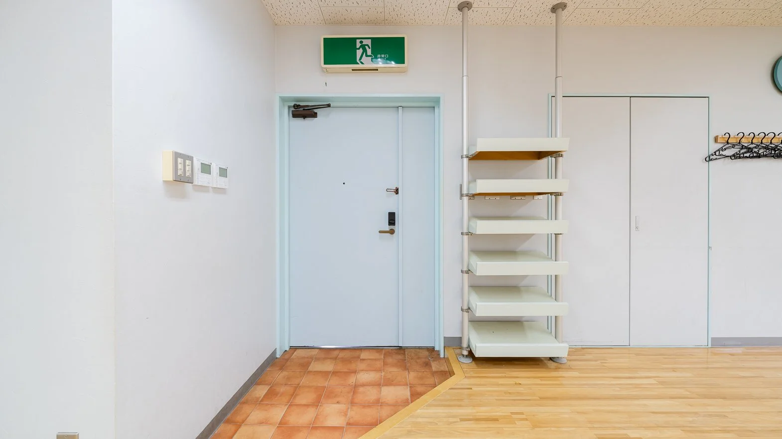 Interior of a room with a white door, green emergency exit sign above, and a coat rack with black hangers on the right. There are shelves on the wall beside the door.