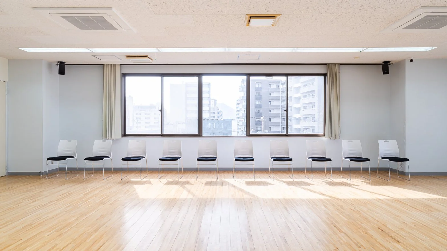 Empty room with a large window, white chairs with black seats arranged in a row, wooden floor, and ceiling vents.