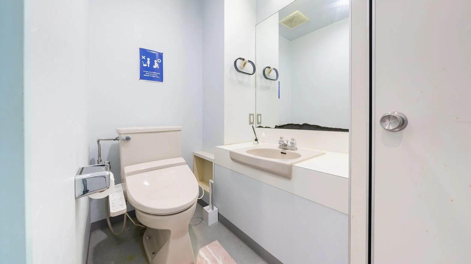 A small bathroom featuring a toilet, a mirror, a sink with a countertop, and a wall-mounted hand towel ring. The walls are plain white, and there is a small sign on the wall above the toilet. The bathroom has a simple, functional design.
