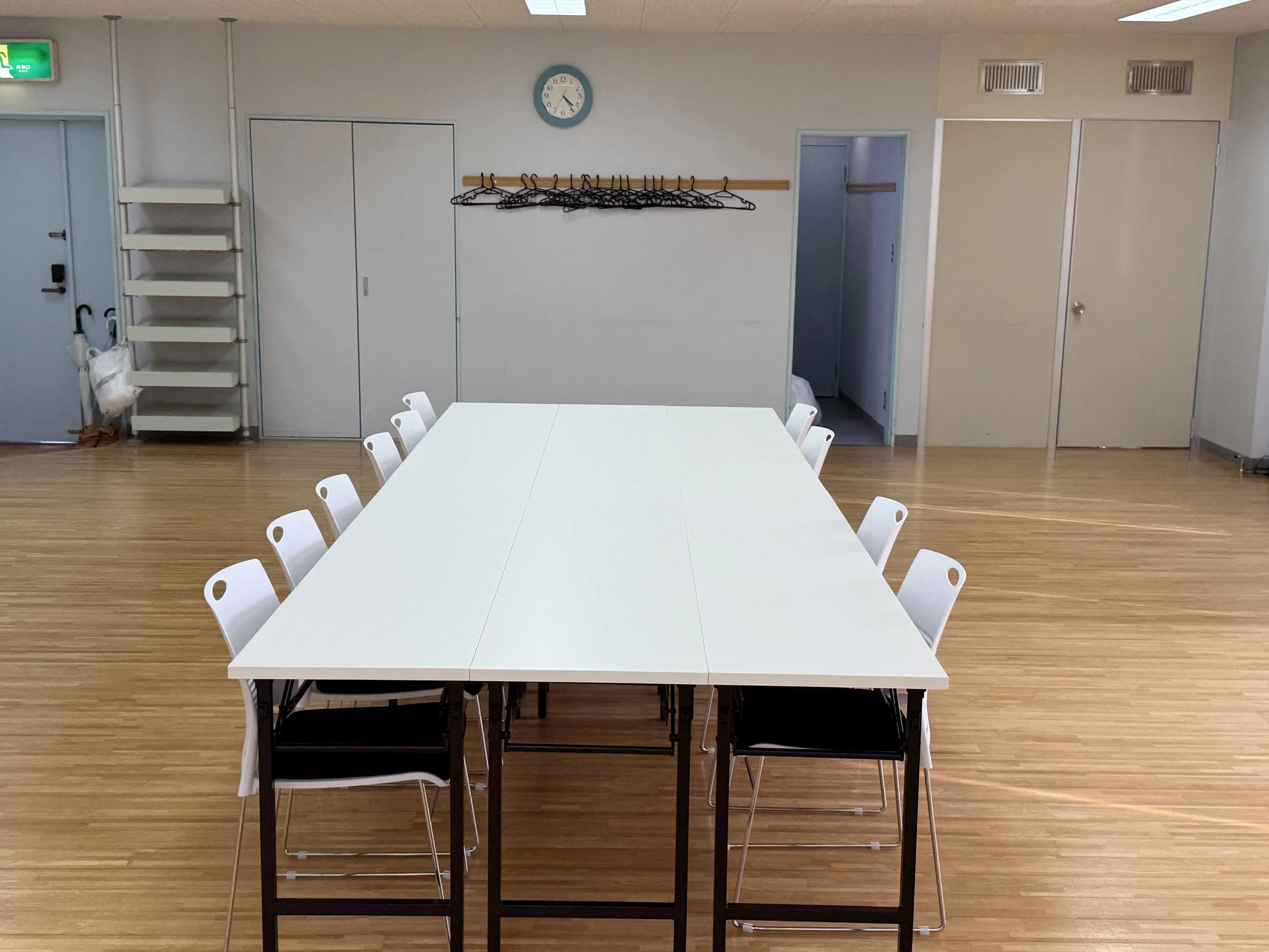 An empty room with a white rectangular table and white chairs, a wall with a clock, coat hangers, and closed doors. There are some shelves with umbrellas near the door, and the room has a wood floor.