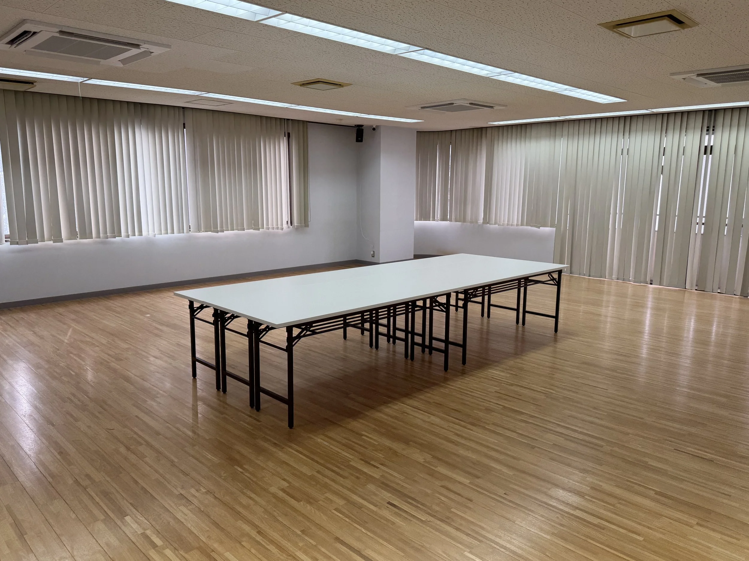 An empty room with large windows covered by vertical blinds, a white table with black metal legs, and wood flooring.