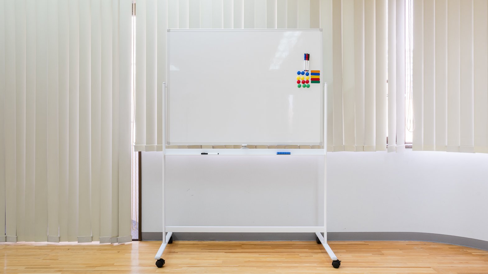 Whiteboard with colorful magnets and markers near vertical blinds in a room with hardwood floor.