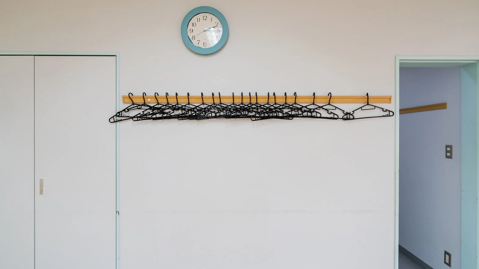 A blue clock with black numbers and hands on a white wall, showing the time as approximately 12:24. Below the clock, there is a wooden rack holding multiple black wire hangers. To the left, there is a closed white door or cabinet, and to the right, an open doorway leading to another room with a white wall and an electrical outlet.