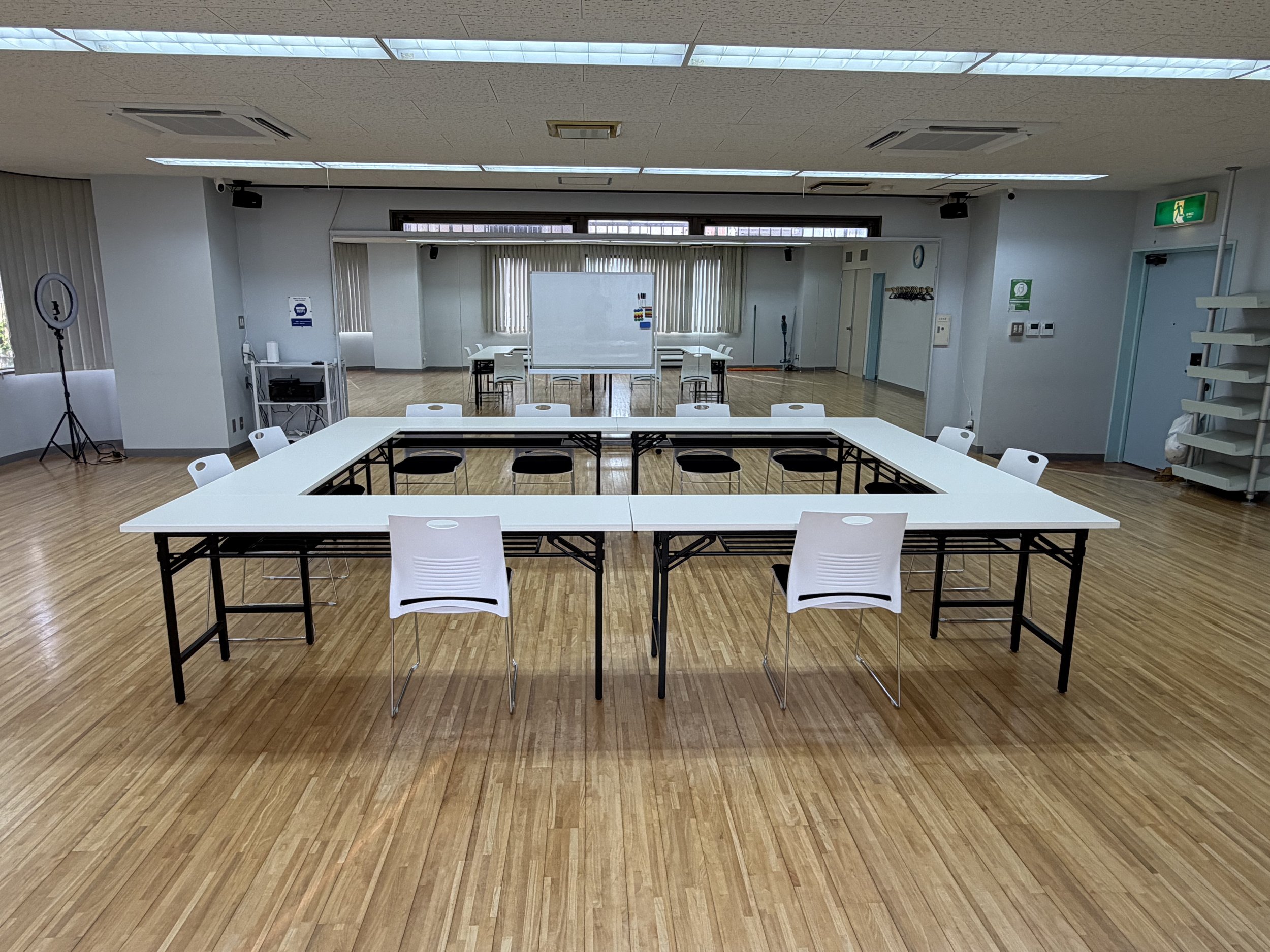An empty conference or meeting room with white tables arranged in a U-shape and white chairs around them. There is a whiteboard and windows with vertical blinds at the back. The room has wooden flooring and ceiling lights.