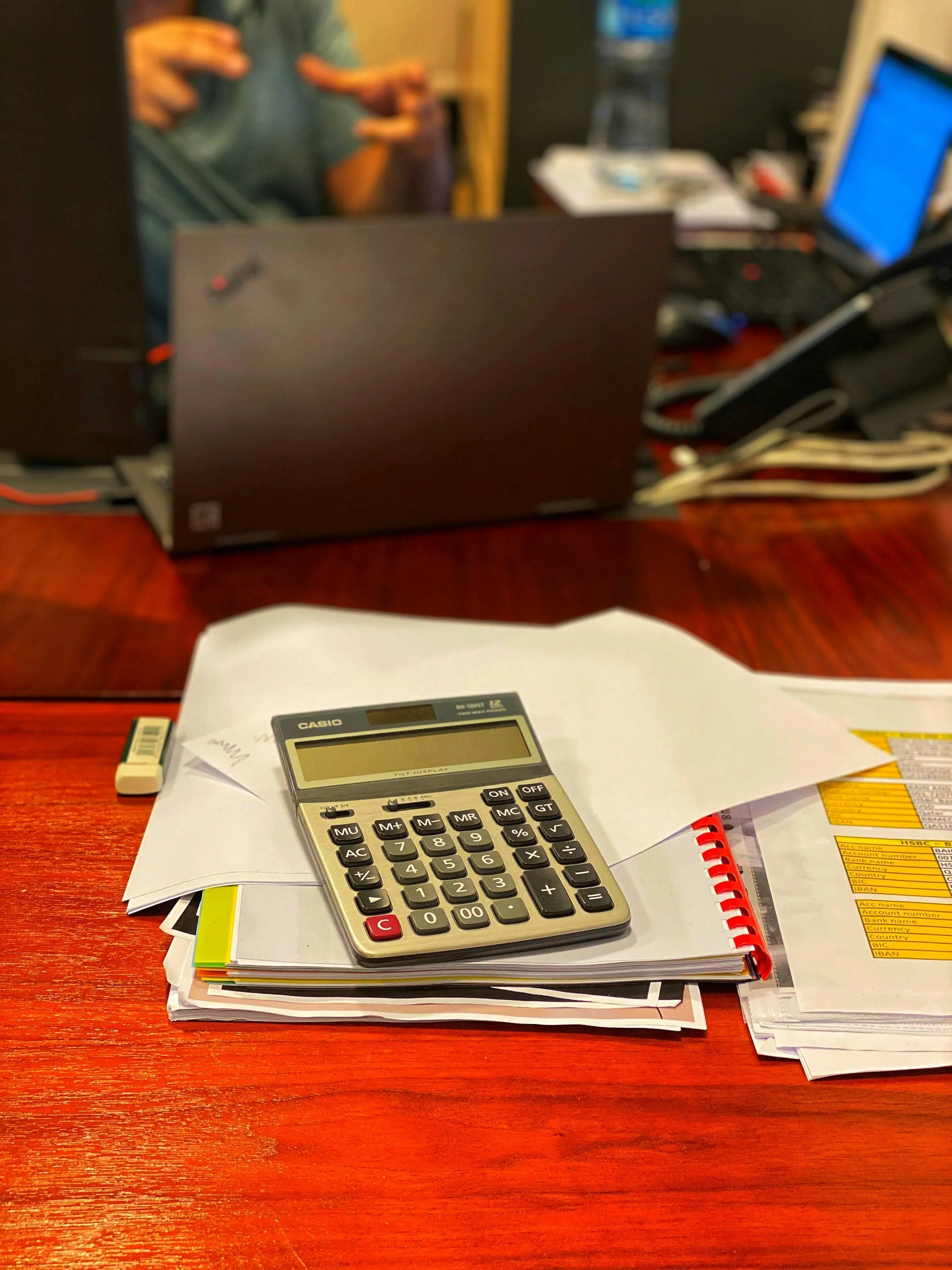Desk with scattered papers, a calculator, a small eraser, and a red spiral notebook, with a blurred person and electronic devices in the background.