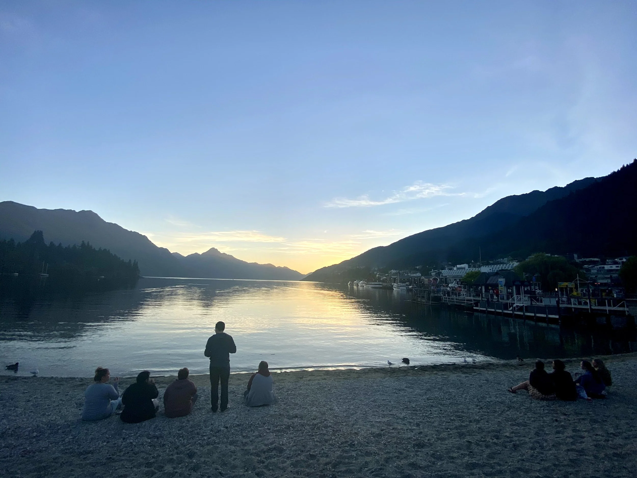 People sitting on a pebble beach near a calm lake at sunset, with mountains in the background and a town along the shoreline.