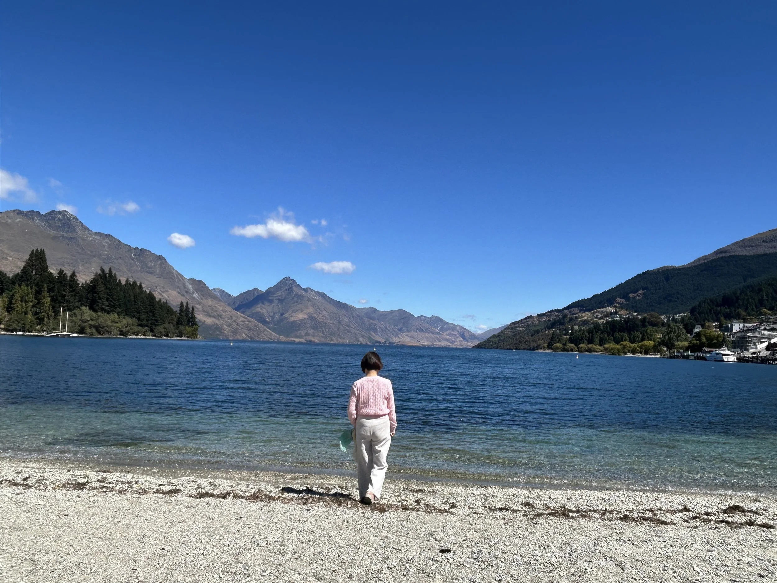 A woman in a pink sweater and white pants walking on a pebbled beach near a large blue lake, surrounded by mountains and a clear blue sky.