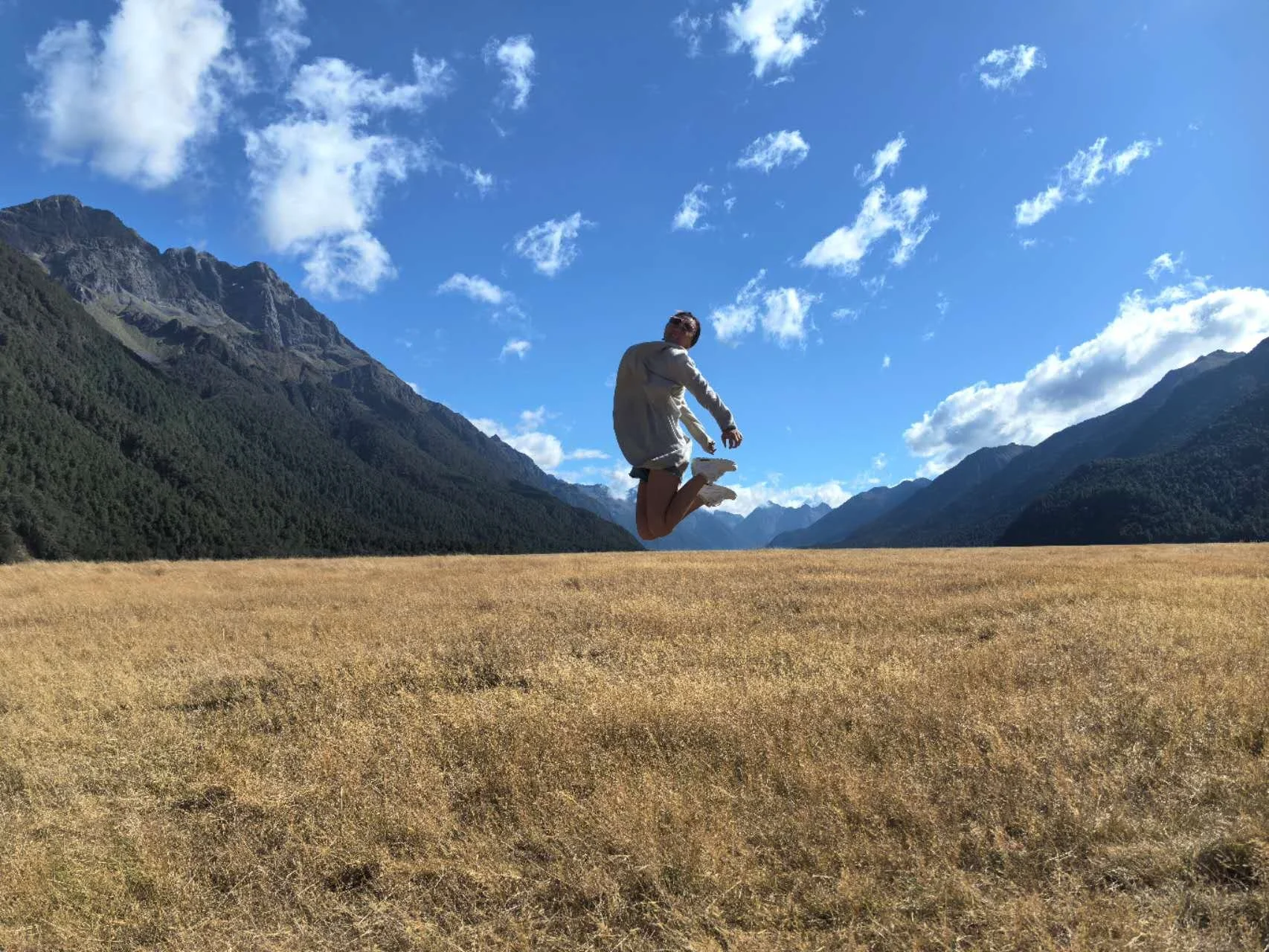 A person mid-jump in a mountainous landscape with a grassy field, forested mountains, and a partly cloudy blue sky.