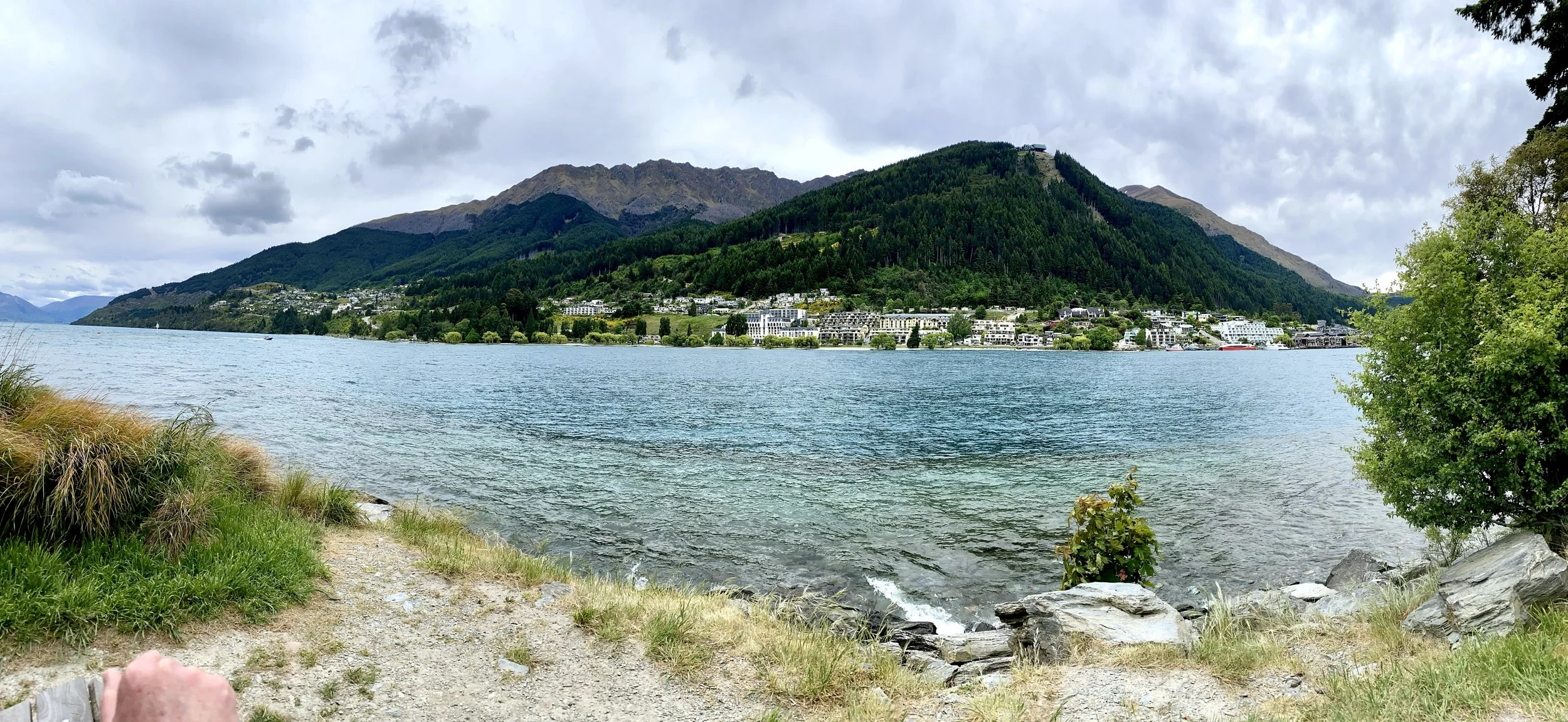 View of a lakeside with a small dirt path, lush grass, and rocks in the foreground. In the background, there are residential buildings at the base of a forested hill or mountain under a cloudy sky.