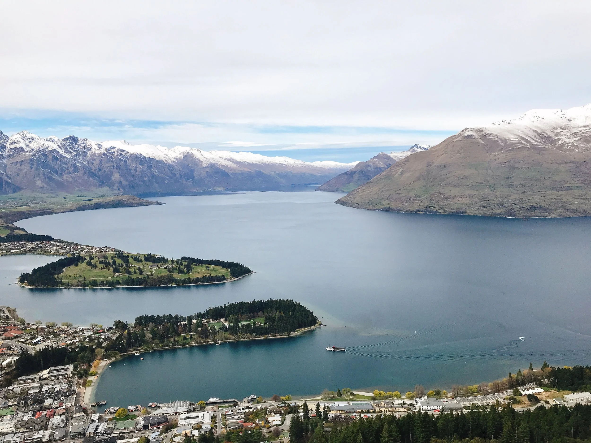 Aerial view of Lake Wakatipu in Queenstown, New Zealand, surrounded by mountains with snow-capped peaks and a small town at the shoreline.
