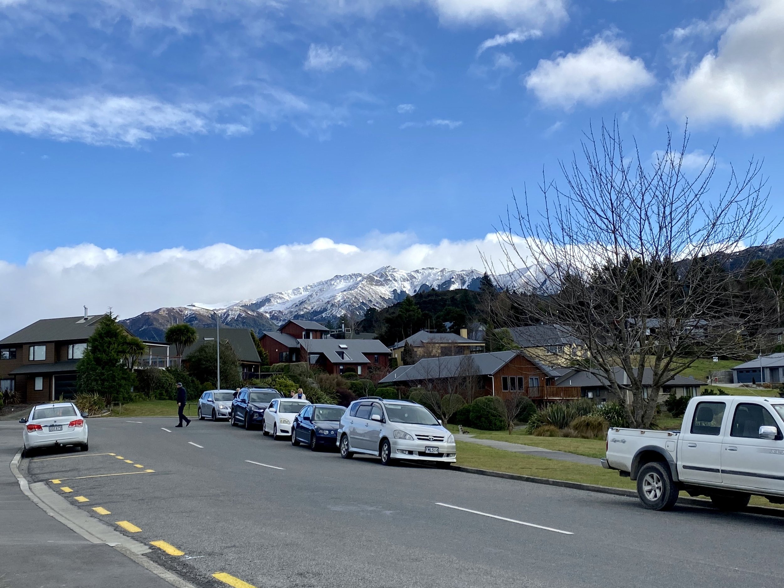 A street view with parked cars, a leafless tree, houses, and snow-capped mountains in the background under a partly cloudy blue sky.
