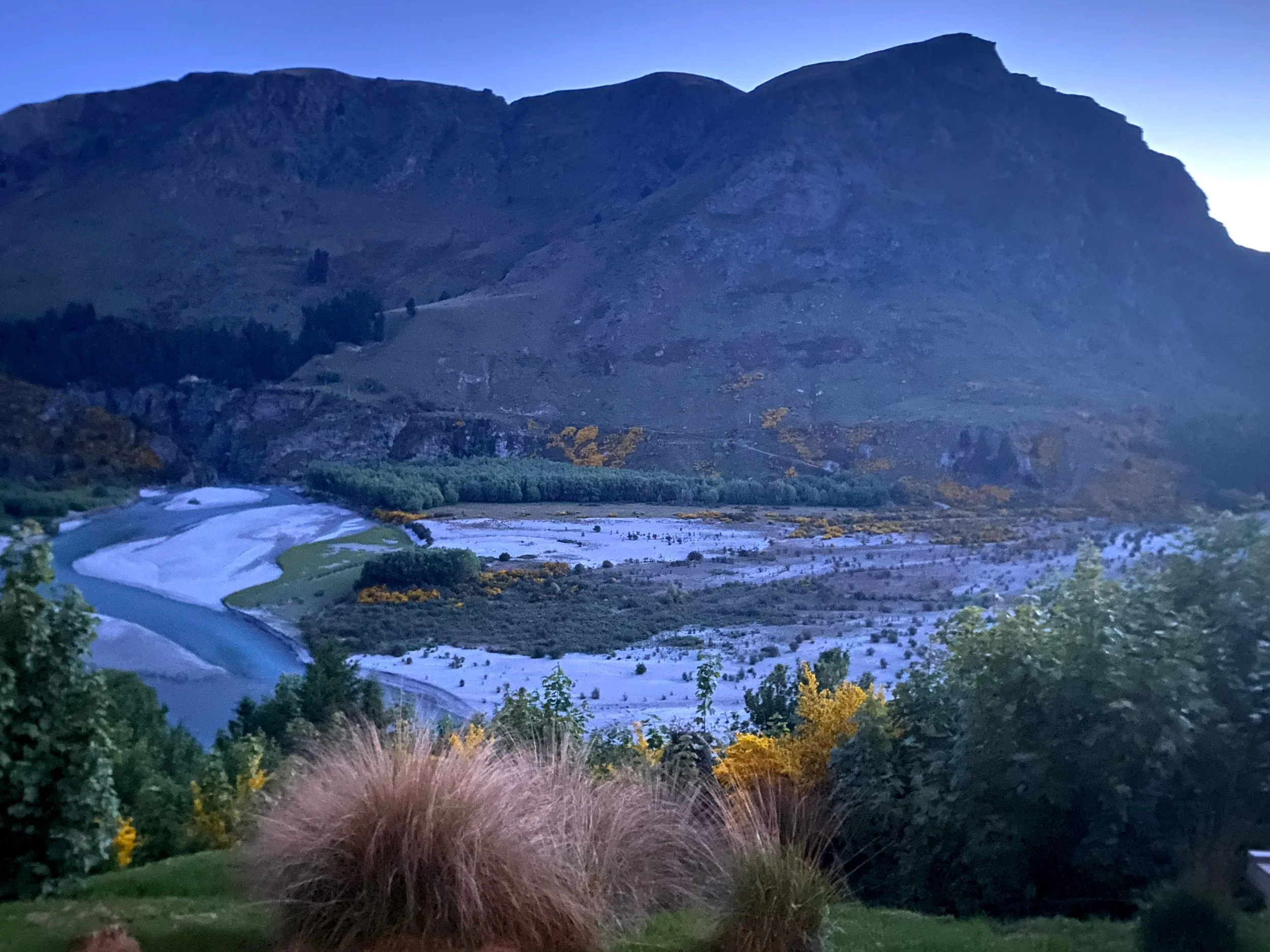 A mountain landscape during dusk with a river winding through a valley surrounded by trees and grassy patches.