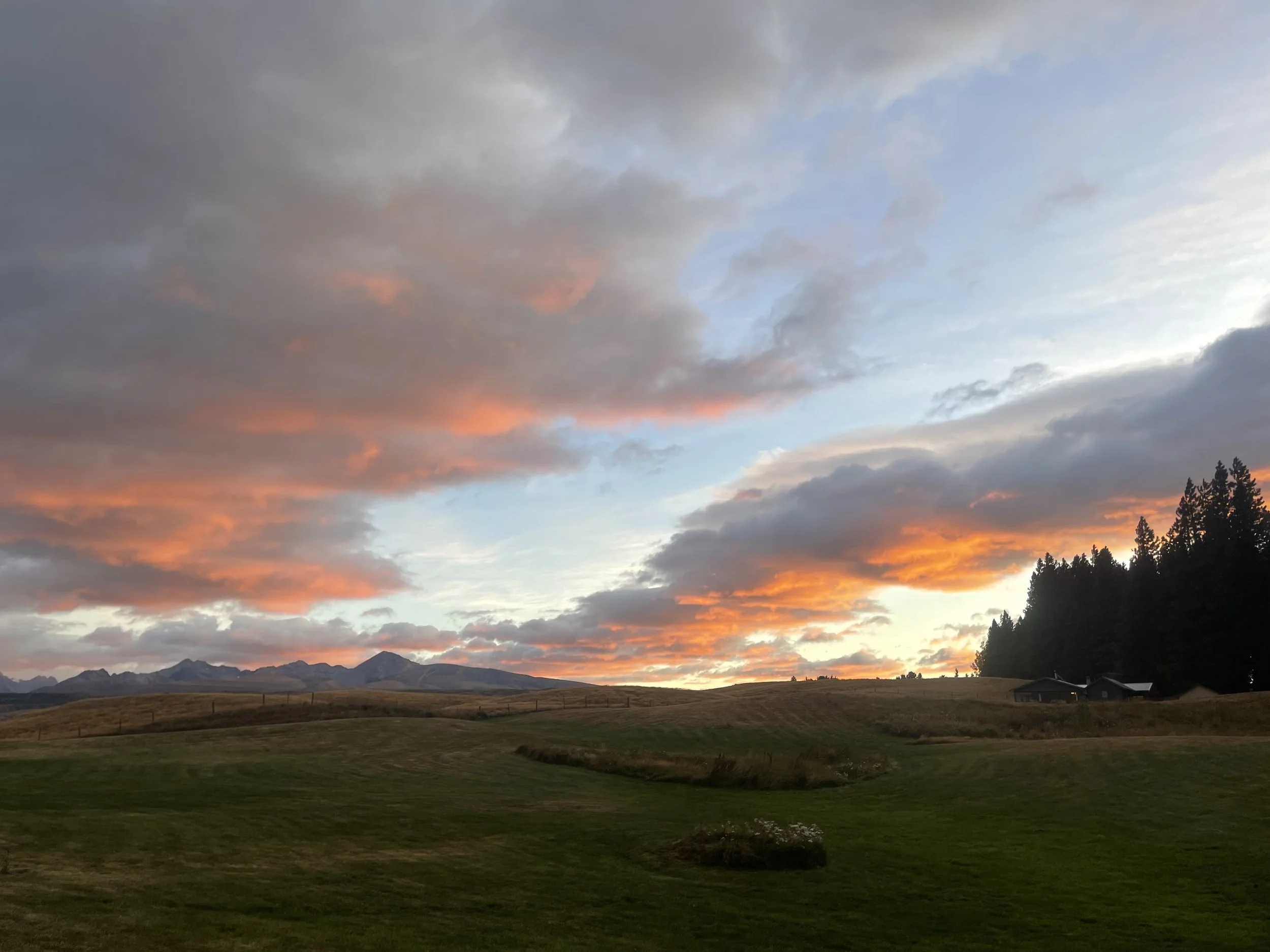 Sunset over rolling green hills, with mountains in the distance and a partly cloudy sky with orange and pink clouds.