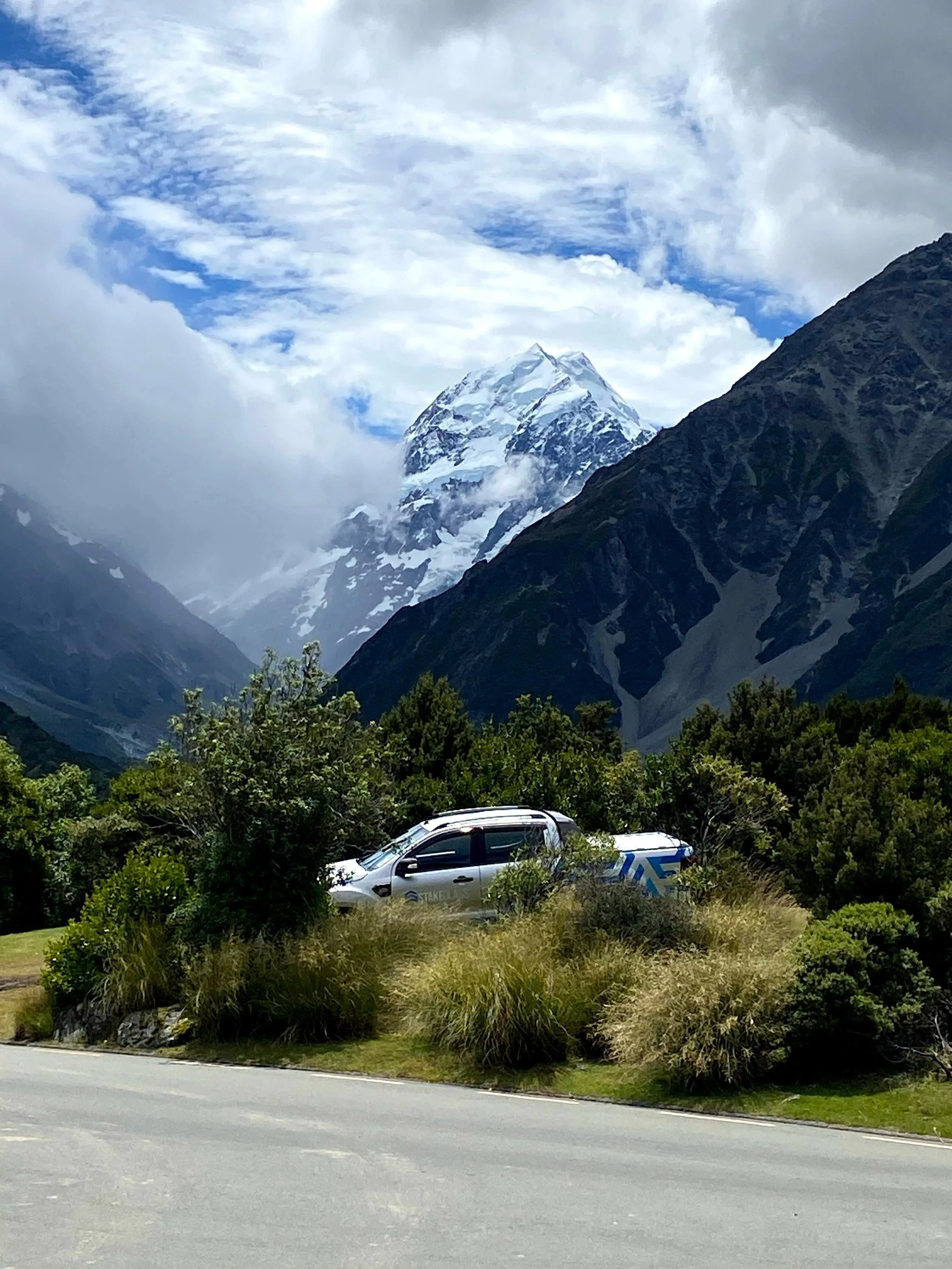A pickup truck parked on a grassy area by the side of a mountain road, with green bushes and trees in the foreground. Snow-capped mountains and a partly cloudy sky are visible in the background.