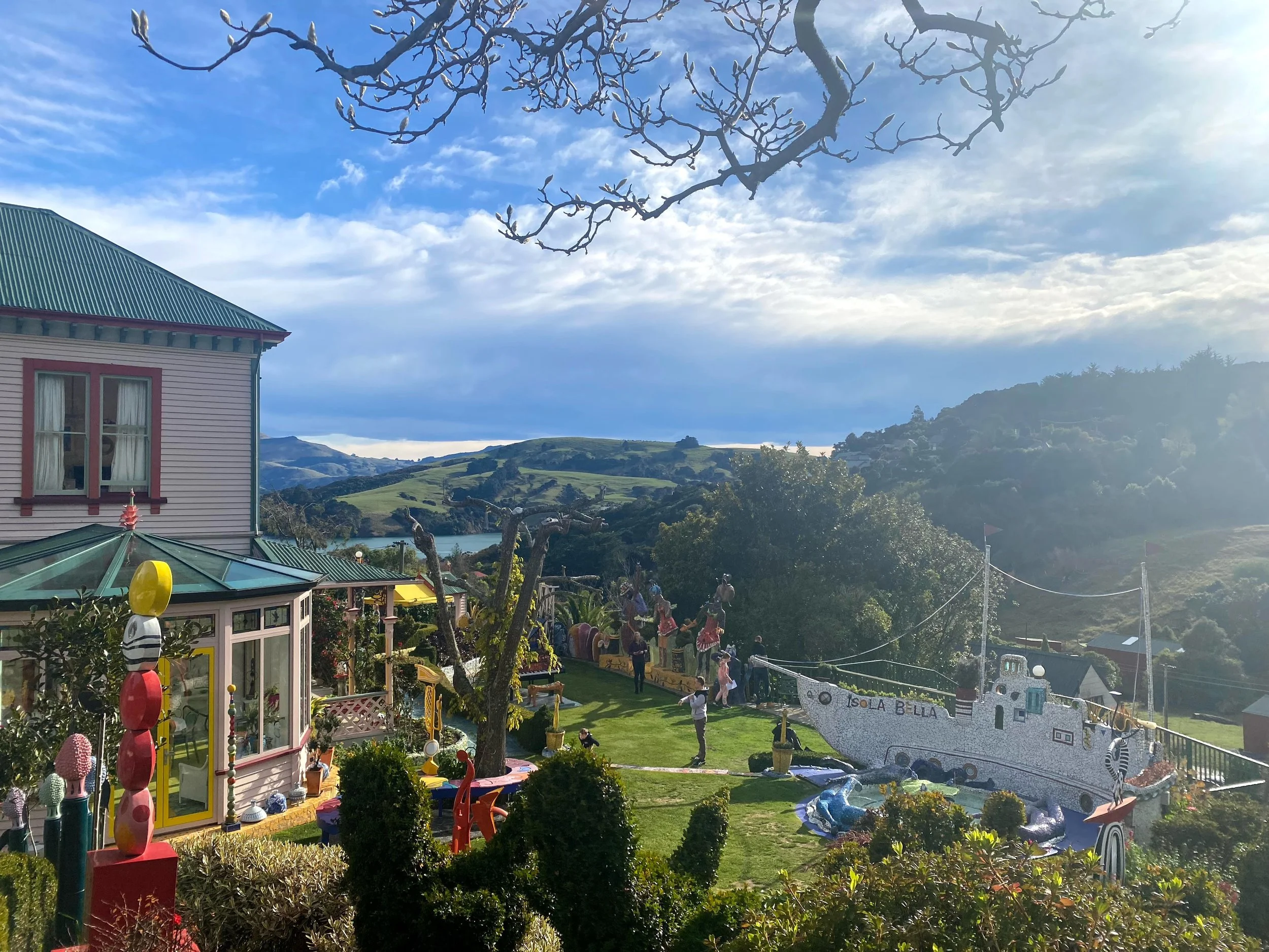 A scenic view of a colorful house with a green roof and large windows, a yard decorated with playful sculptures including a ship named Iso B. Ella, and a beautiful landscape of rolling hills and a lake in the background under a partly cloudy sky.