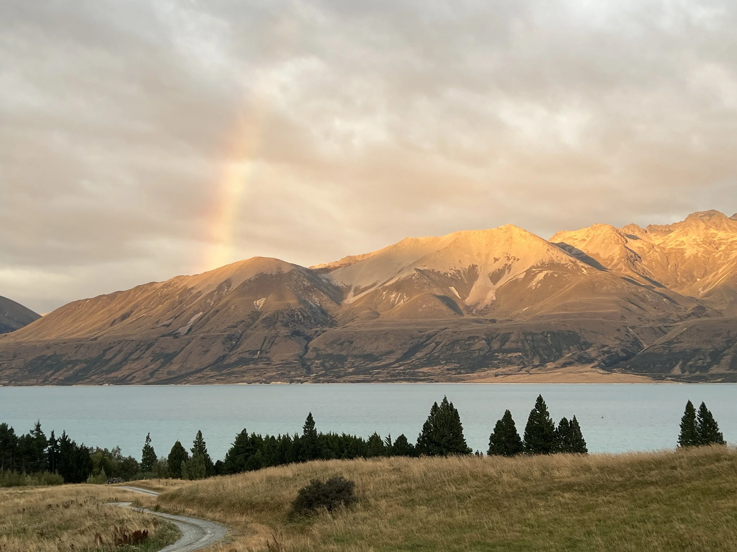Scenic landscape of a mountain range with snow-capped peaks illuminated by sunlight, a calm teal lake in the foreground, and a grassy field with trees along the shoreline. A faint rainbow is visible in the cloudy sky.