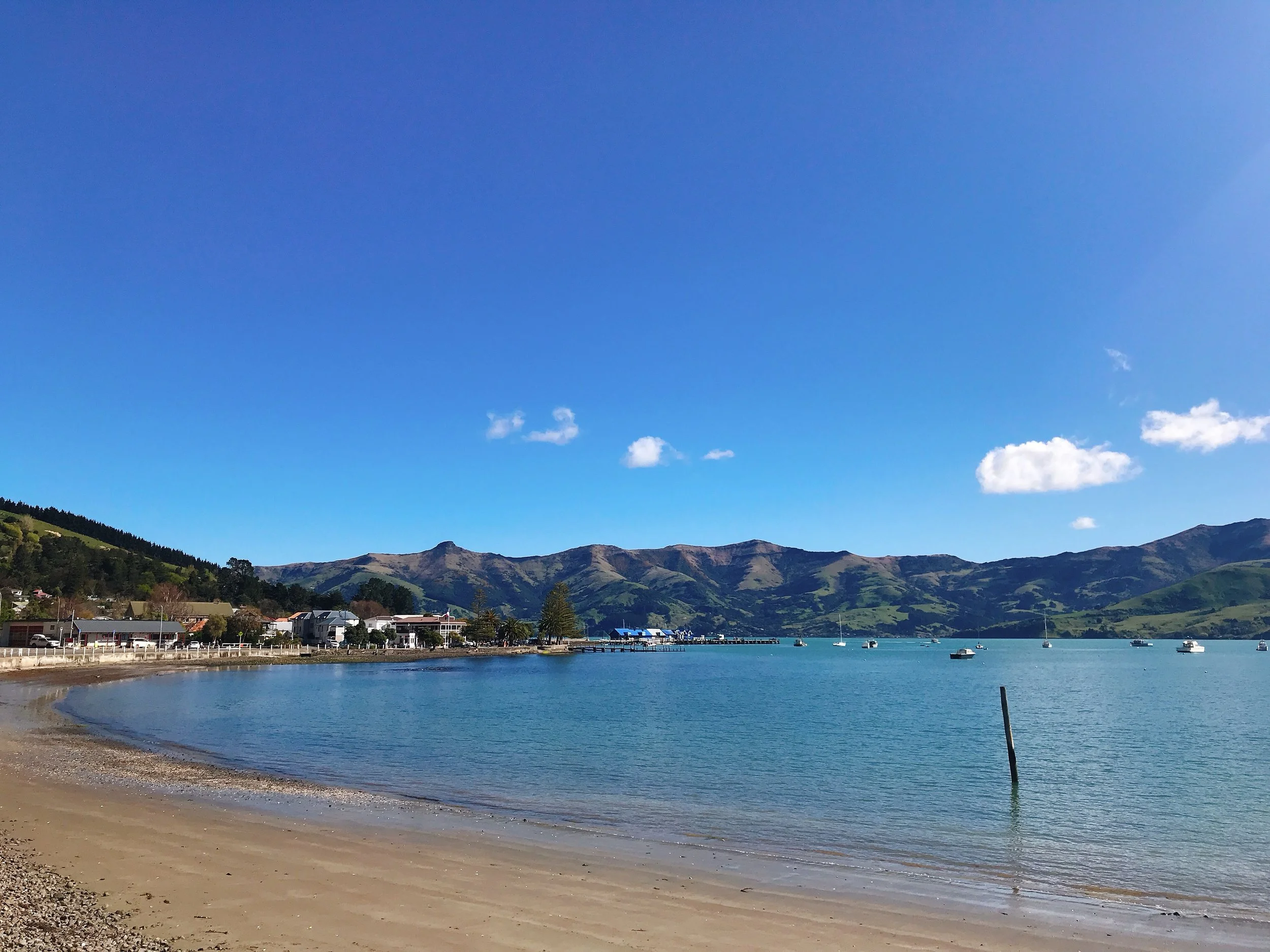 Sunny day at a small coastal town with a sandy beach, calm blue water, and mountains in the background. Small boats are anchored in the water near a pier. There are a few clouds in the clear sky.