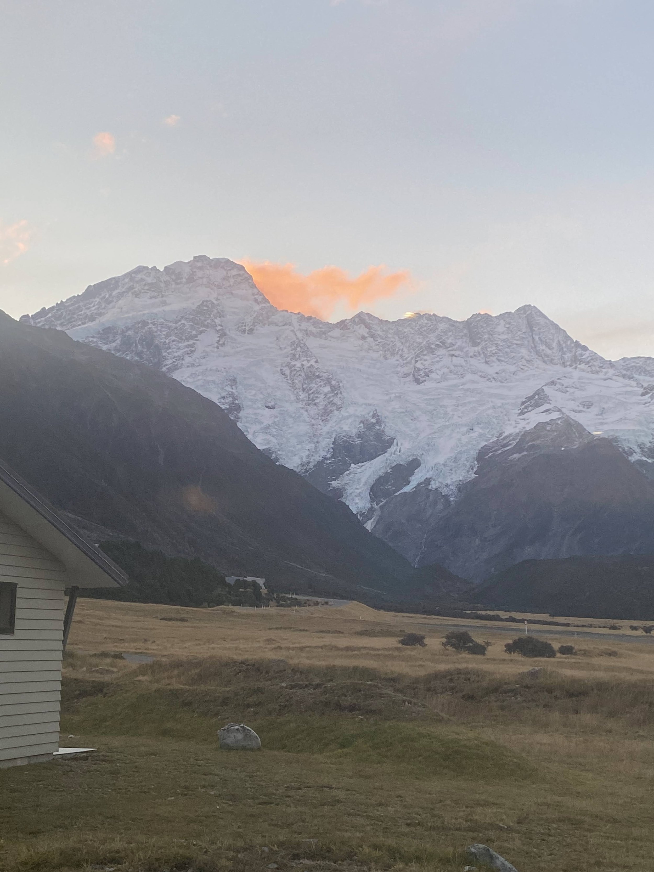 Snow-capped mountains with a pink cloud at sunset, a mountain house on the left, and a grassy field in the foreground.