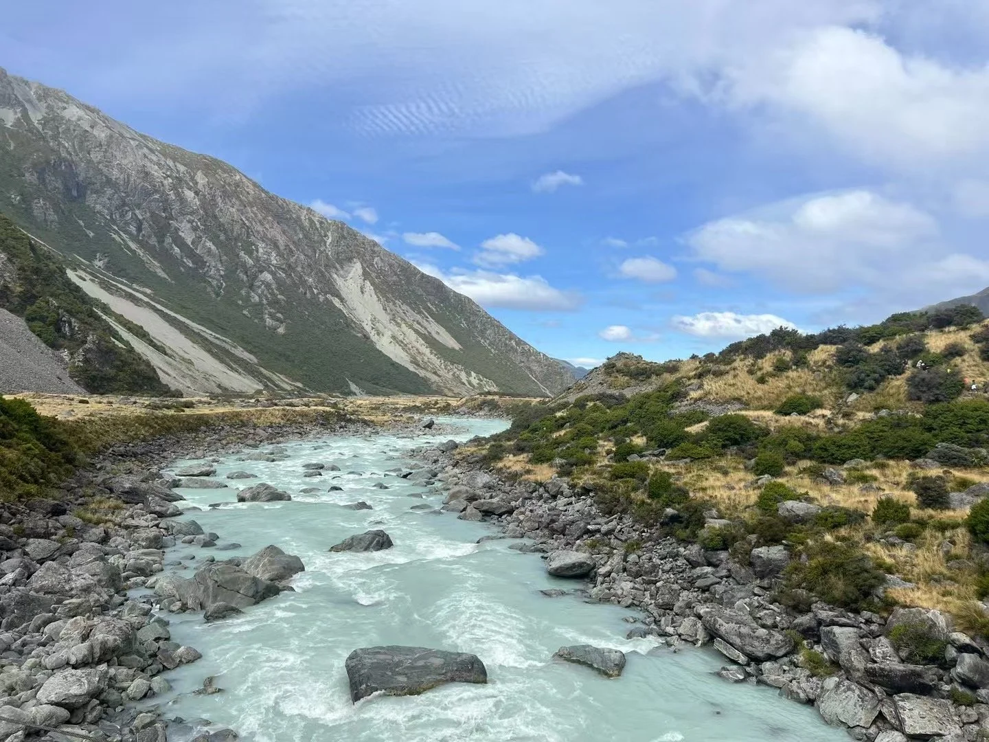 A river flows through a rocky valley surrounded by mountains with green vegetation, under a partly cloudy sky.