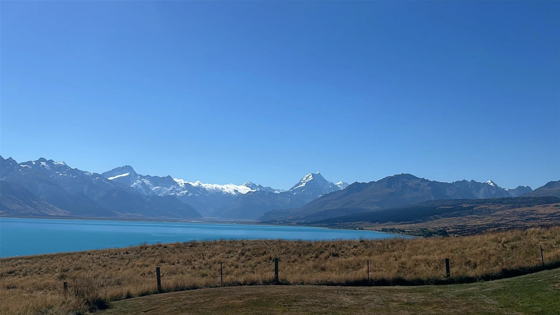 Scenic view of a large lake surrounded by grass and rolling hills, with snow-capped mountains in the distance under a clear blue sky.