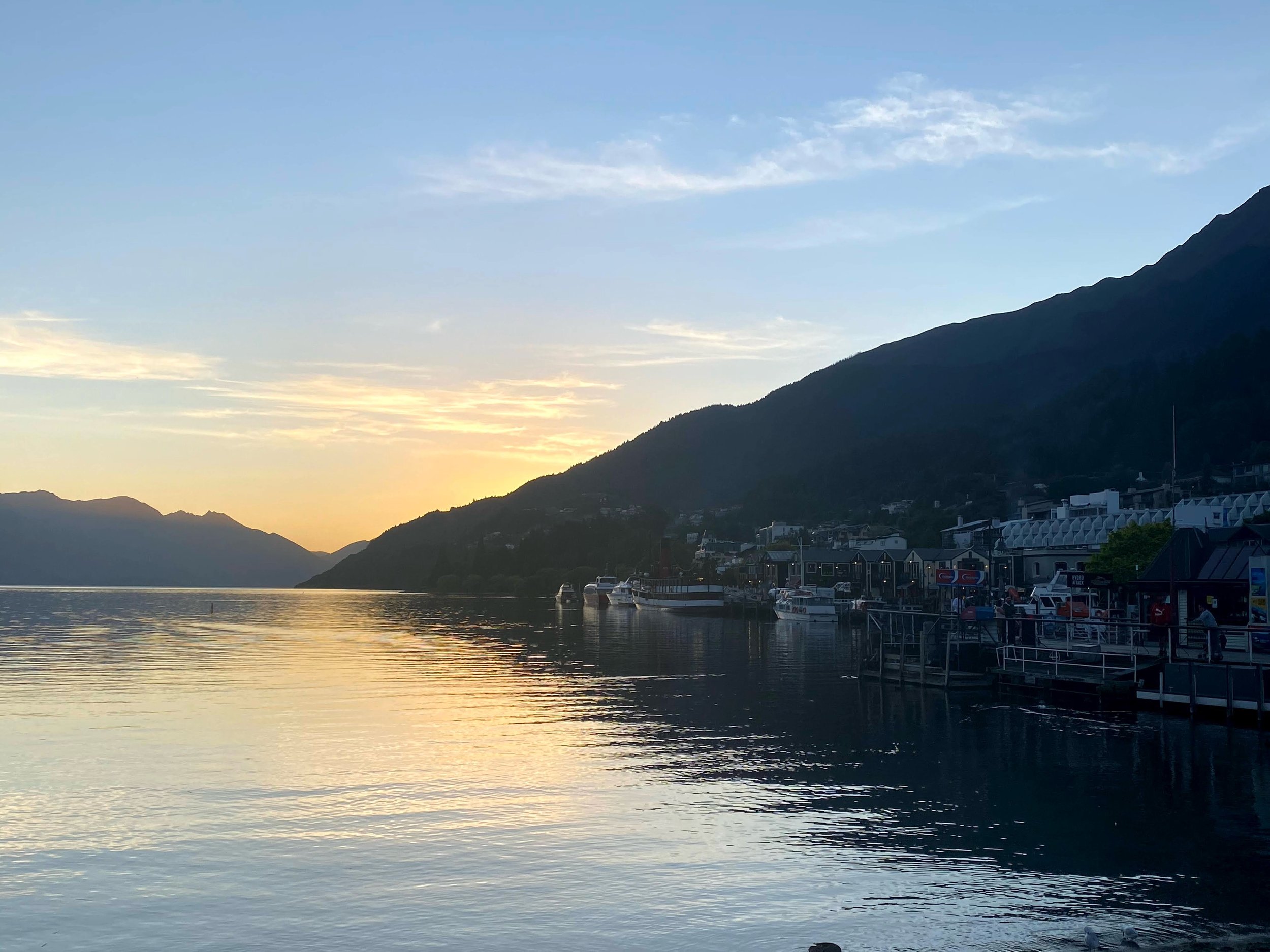 Sunset over a tranquil lake with mountains in the background, a small dock with boats and buildings along the shoreline.