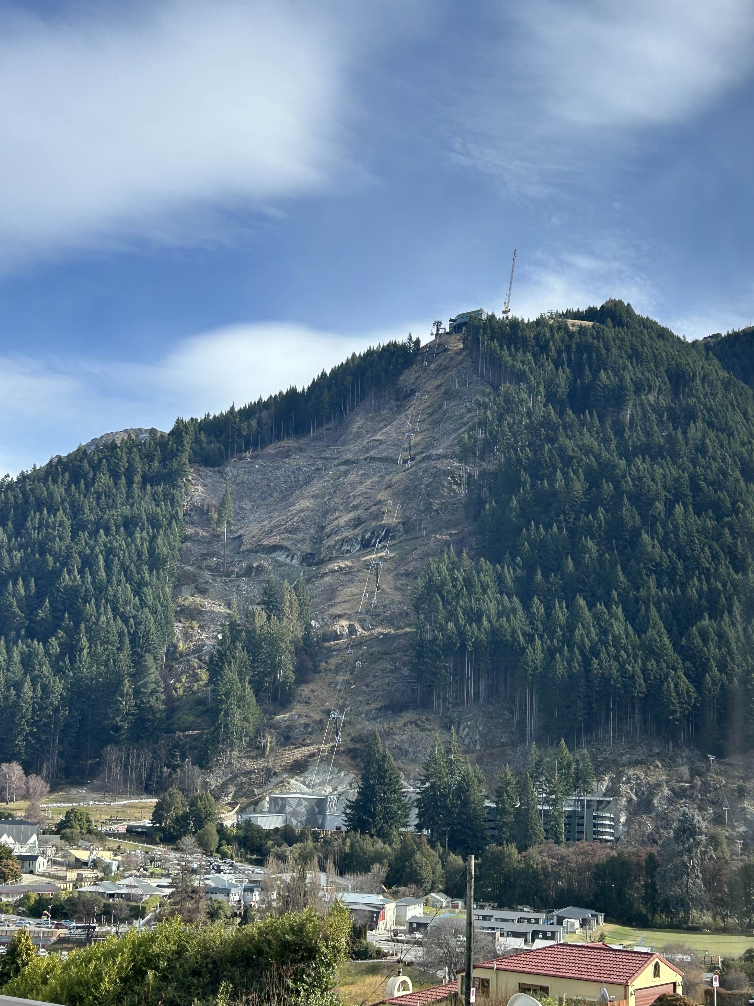 A mountain with dense green trees and a ski lift system running up its slope. The sky is partly cloudy.