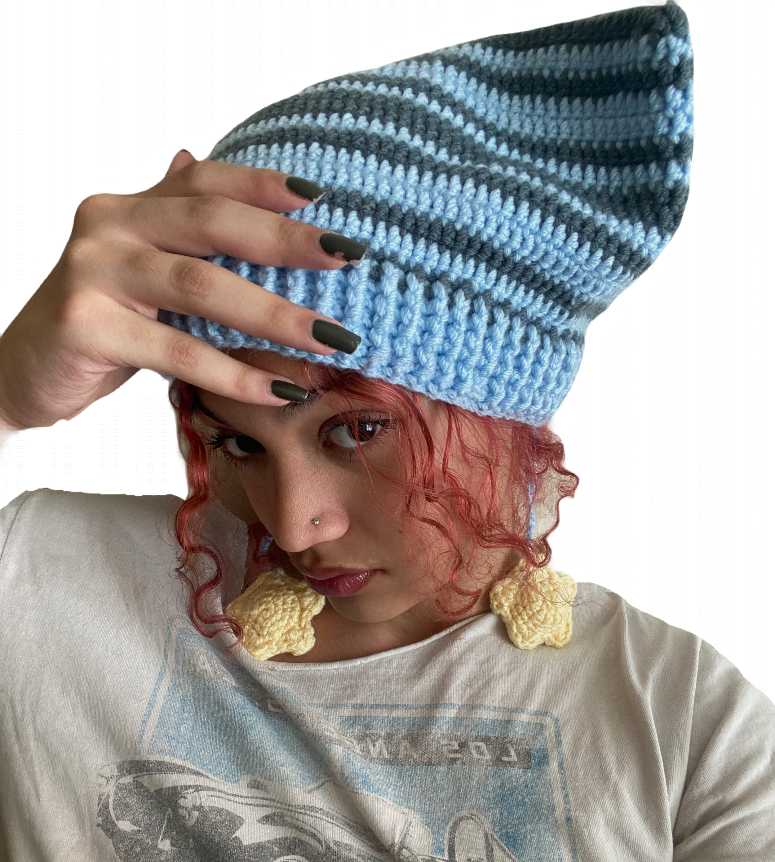 A woman with red curly hair and a nose piercing wears a beige t-shirt, large yellow crocheted earrings, and a blue and gray striped knit beanie. She holds her hand with dark nail polish to the beanie, partially covering her forehead, while looking into the camera.