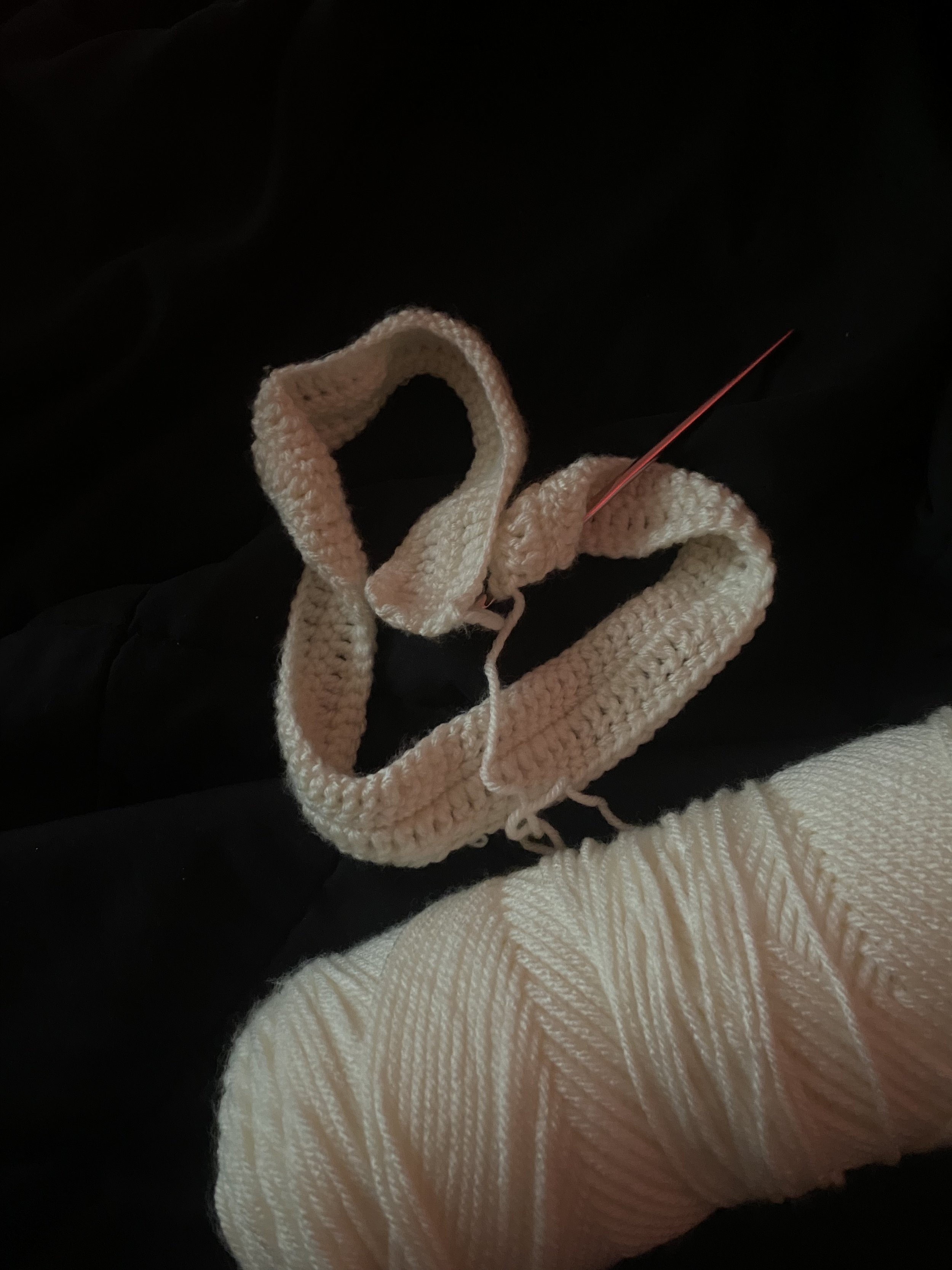 A ball of off-white yarn and a partially completed knitted or crocheted piece in off-white yarn, with a knitting needle inserted, on a dark background.