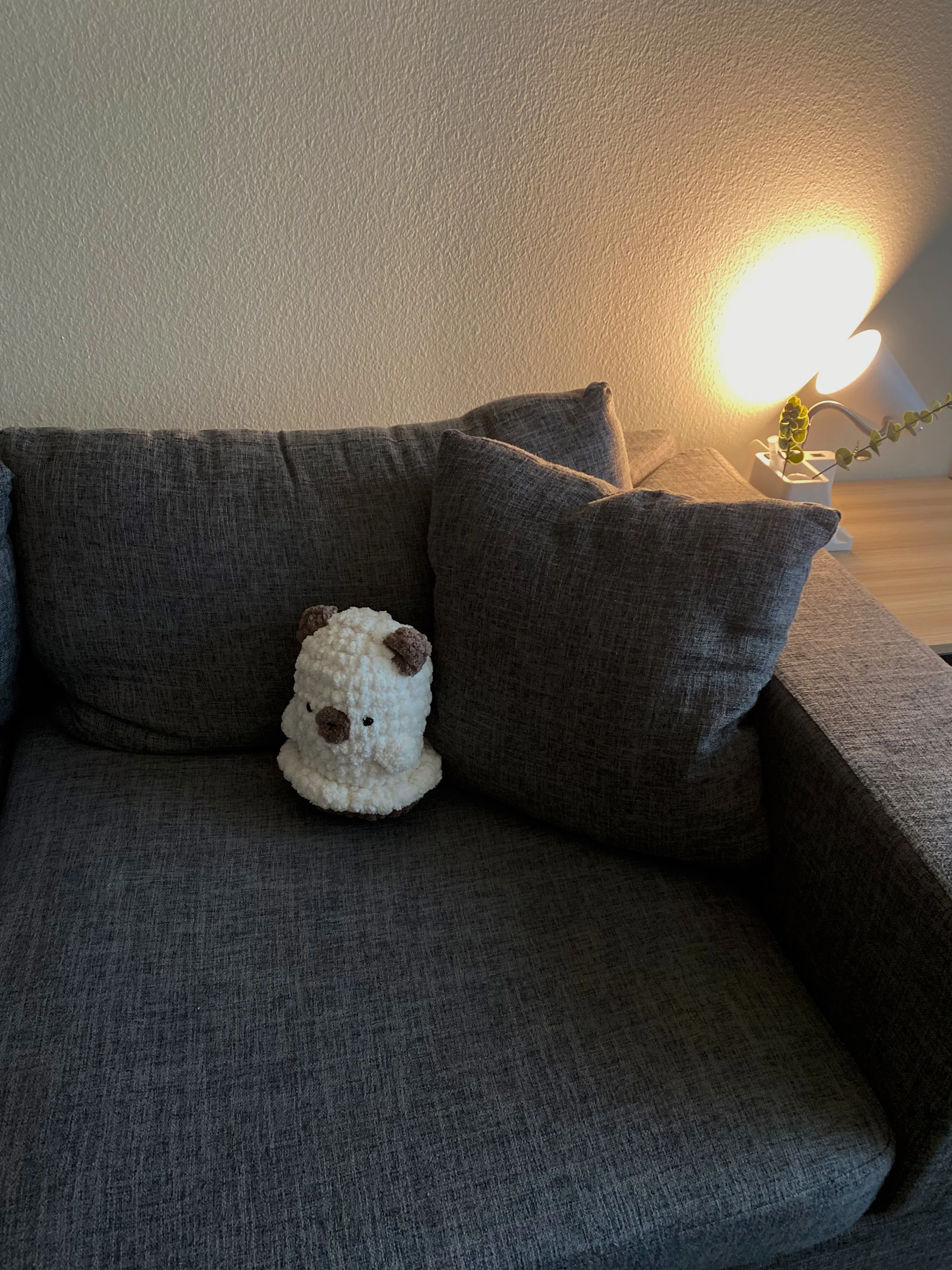 Gray upholstered sofa with dark gray cushions, a plush animal toy resembling a bear or dog, a corner lamp casting warm light, and a small potted plant on a wooden side table.