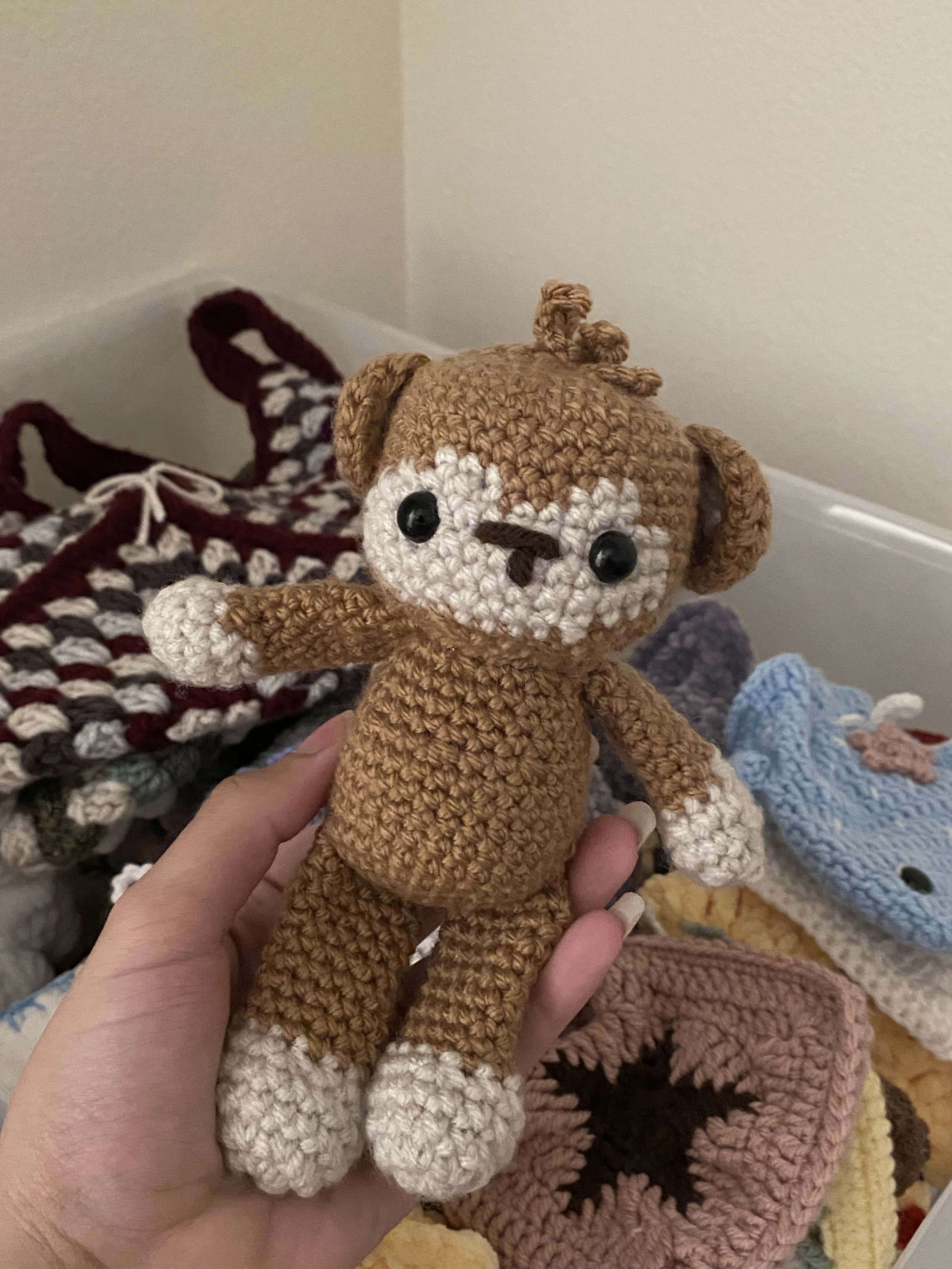 Hand holding a crocheted teddy bear with brown body, white face, paws, feet, black eyes, and a little nose, with crochet blankets and other handmade items in the background.