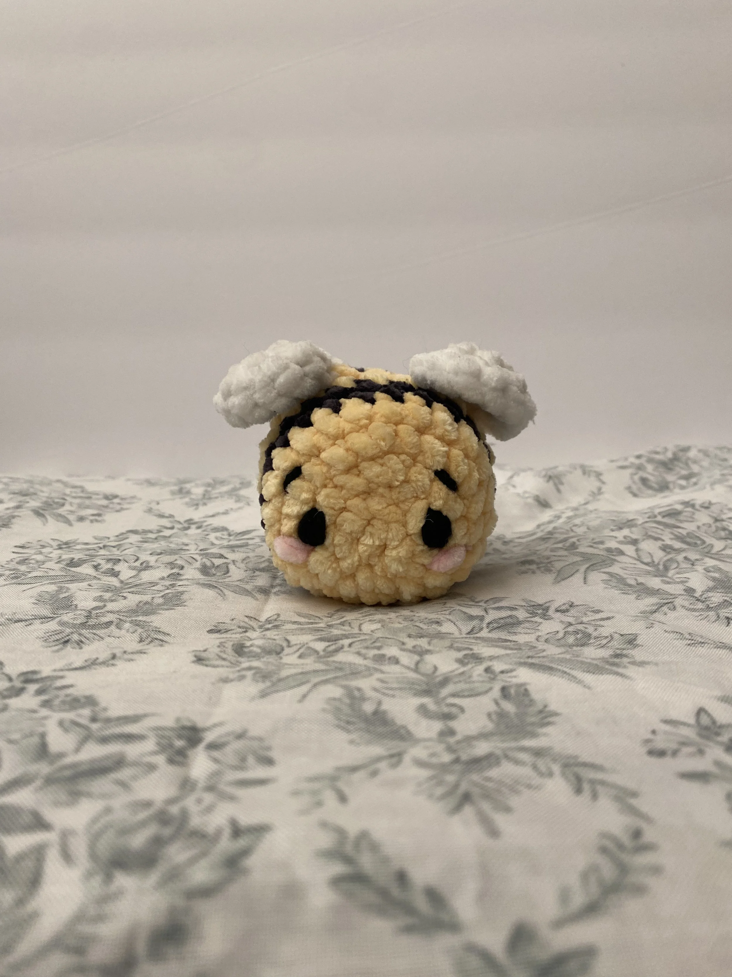 Close-up of a small, plush, handmade toy resembling a cute animal with a round, yellow body, black eyes, pink cheeks, and small white ears, placed on a patterned fabric surface with a plain background.