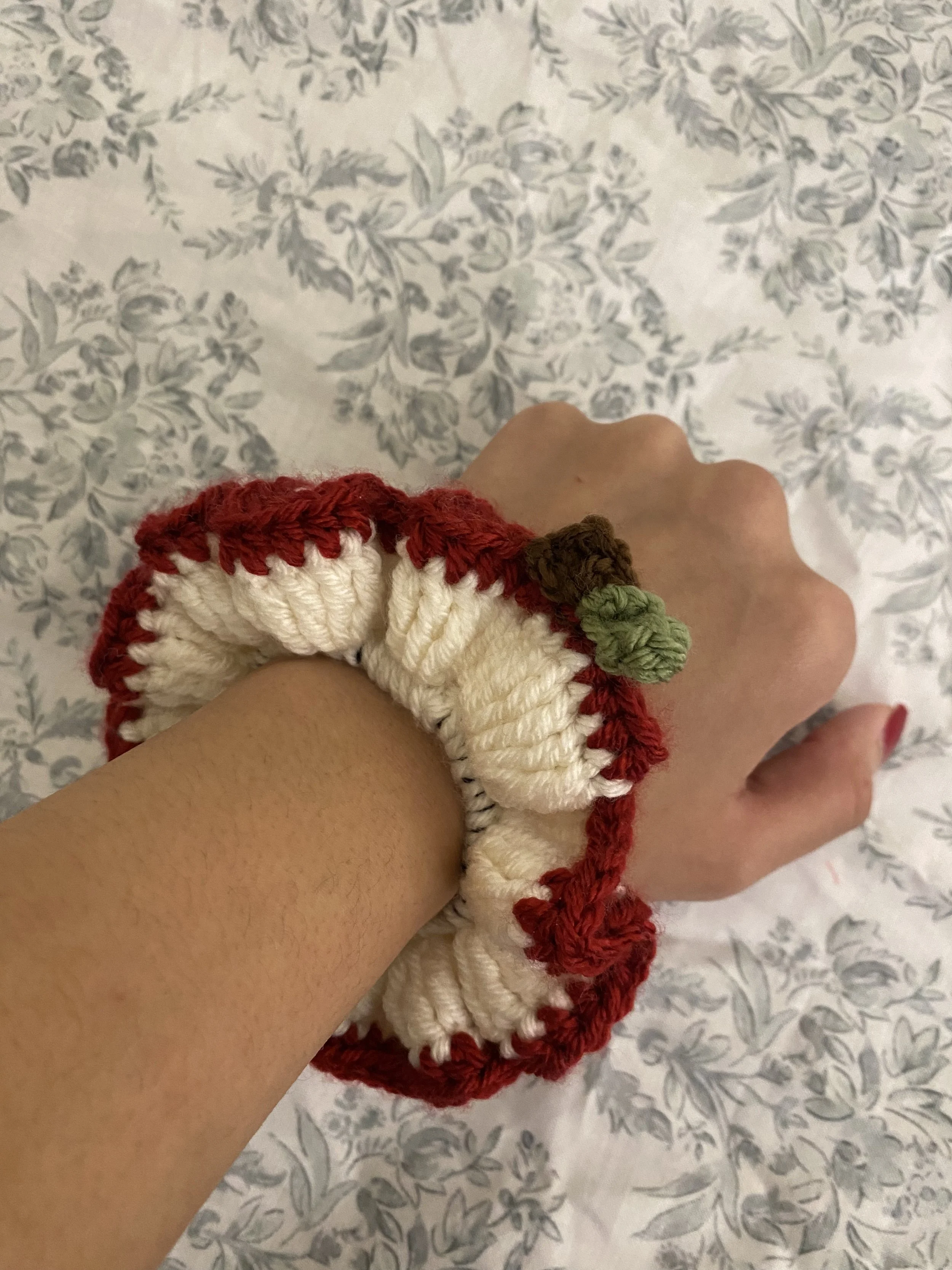 A person's hand and wrist with a knitted wristband that resembles a ladybug. The wristband has a red and white body with a green appendage, set against a background of a floral-patterned fabric.