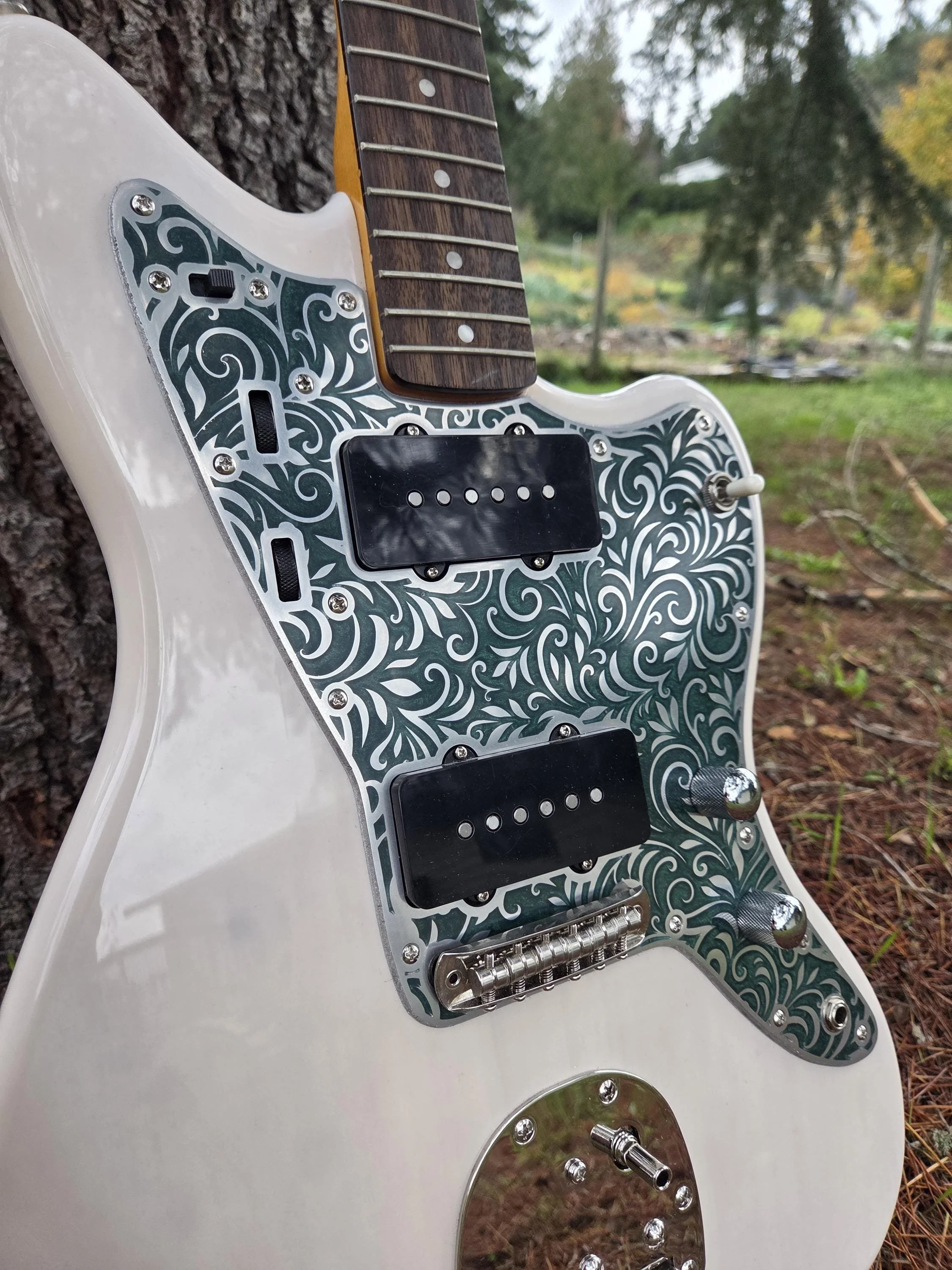 An electric guitar with a white body, ornate metallic pickguard, two black pickups, and knobs and switches, leaning against a tree outdoors.