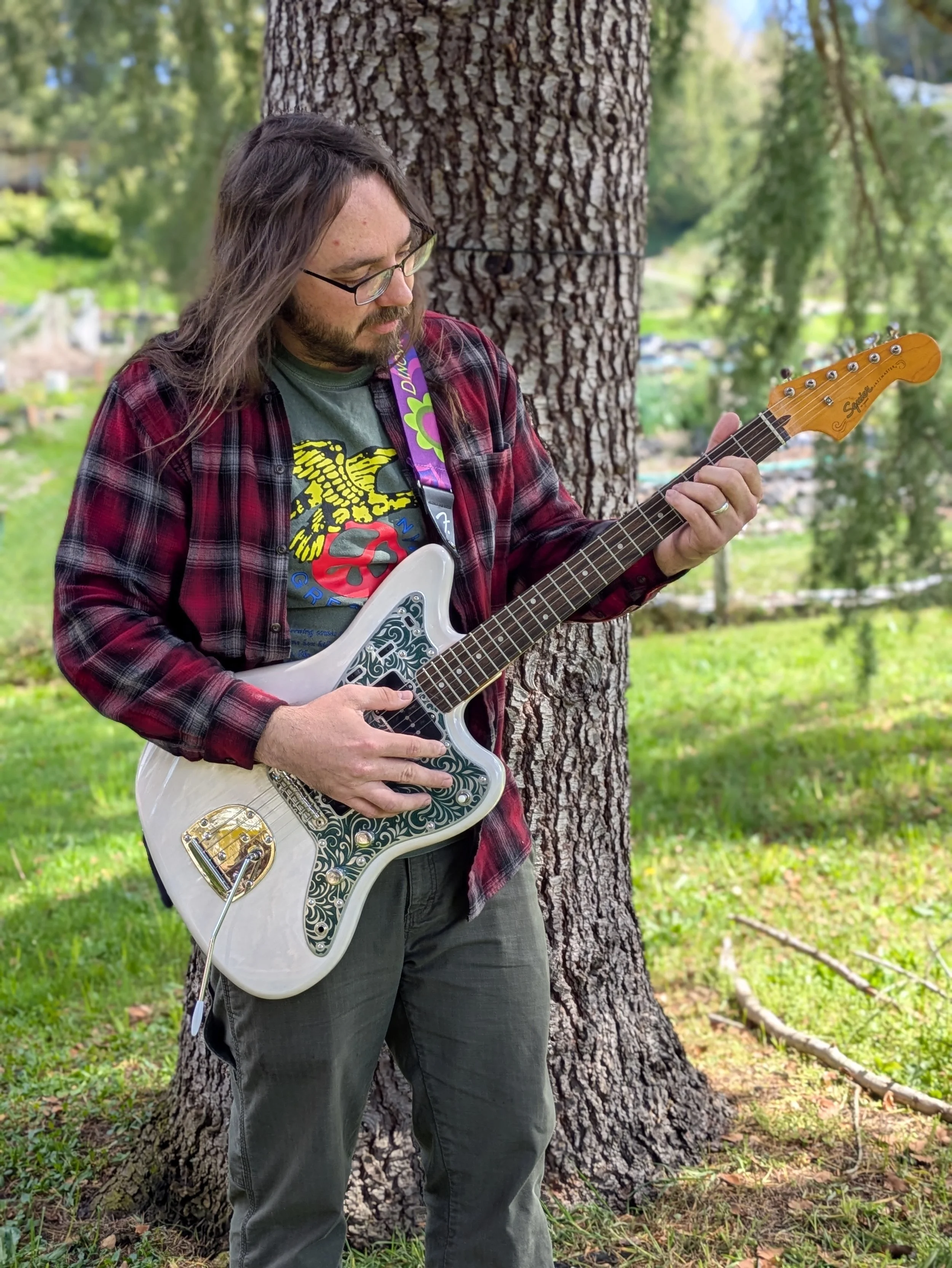 A man with long hair and glasses playing an electric guitar outdoors next to a large tree, with a grassy area and trees in the background.