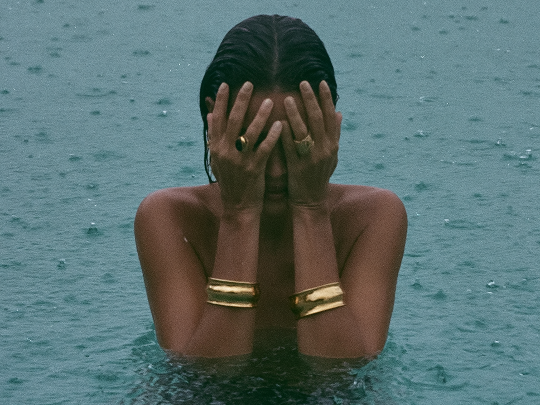 Woman standing chest-deep in open water during rainfall, hands covering her face, wearing gold cuff bracelets and rings, wet dark hair slicked back.