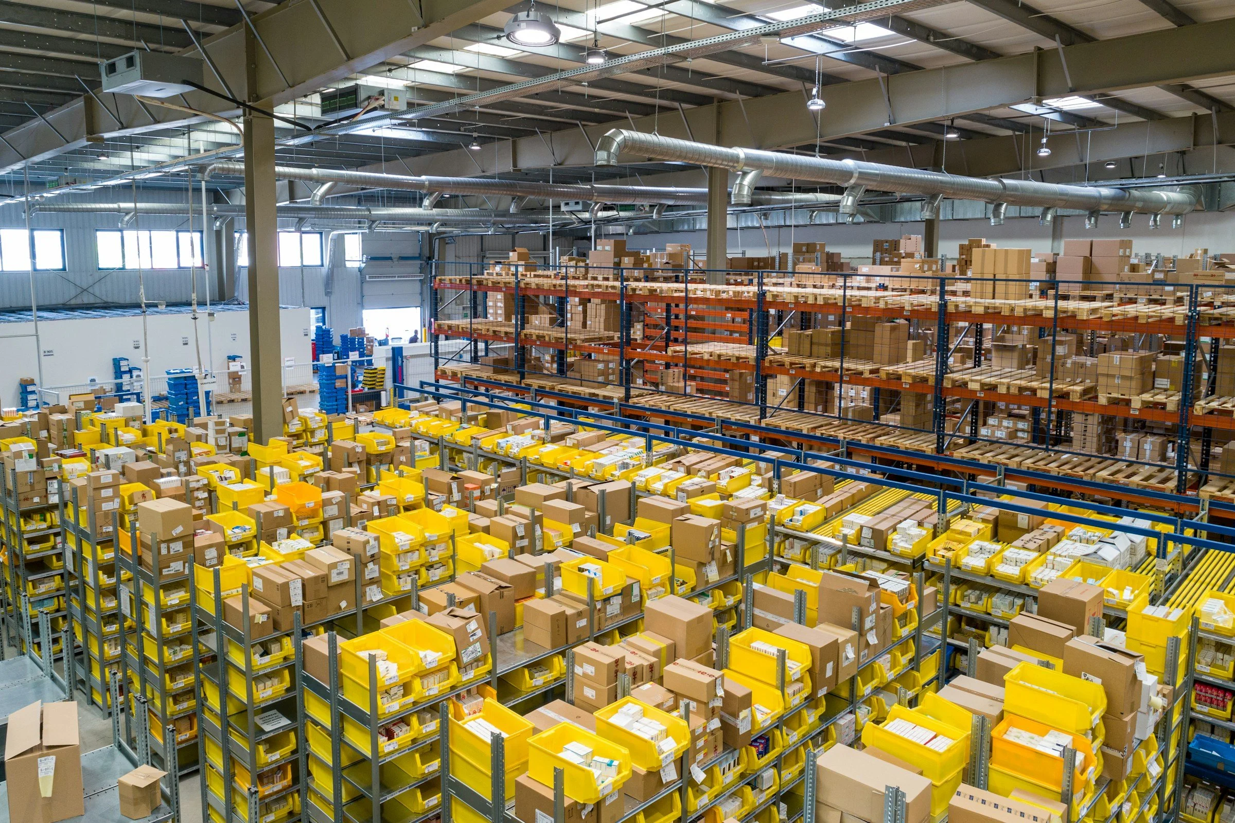 A large warehouse with high ceilings, filled with shelves and boxes, some in yellow bins, and high industrial lighting.