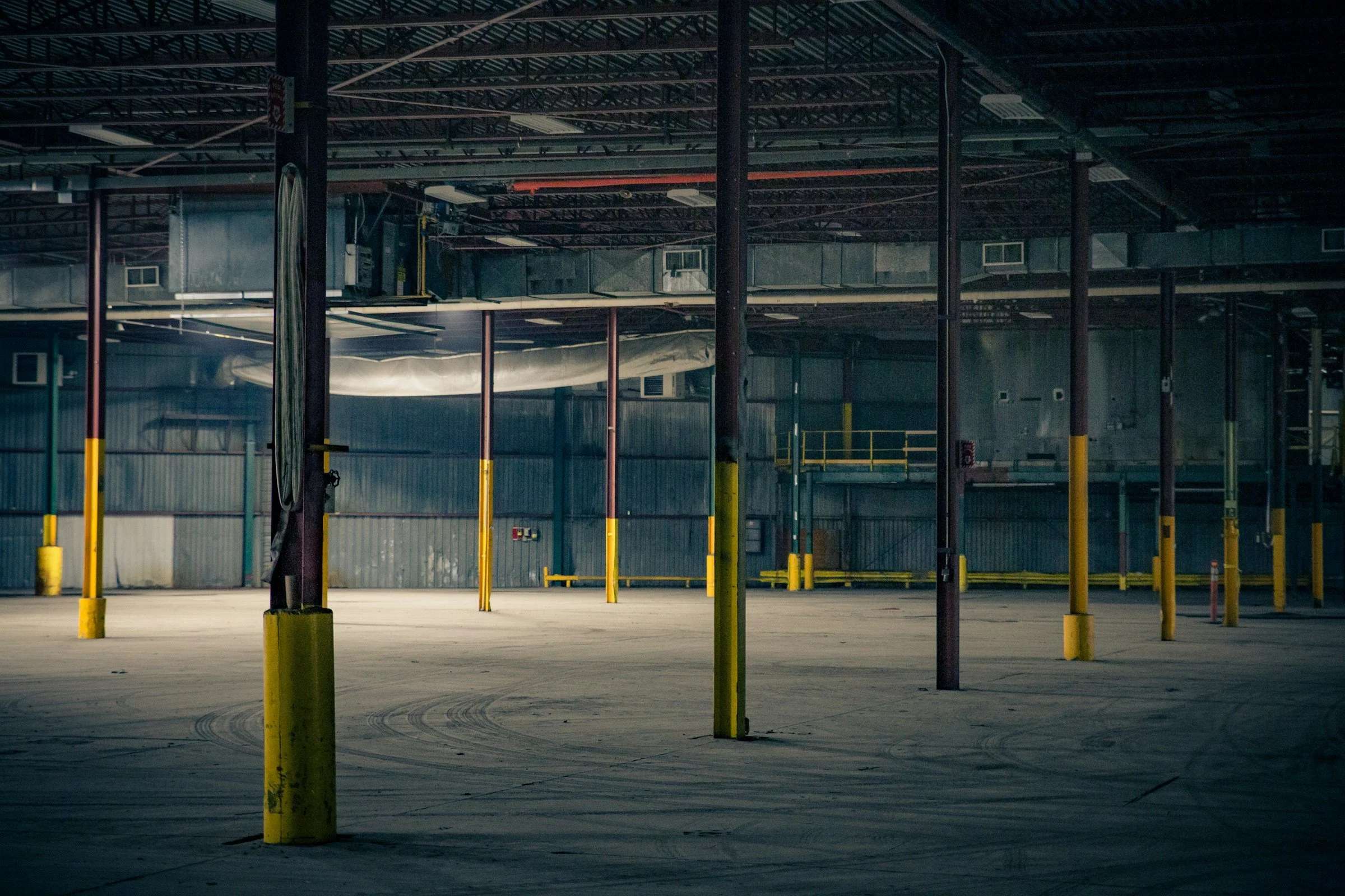 Empty industrial warehouse with yellow safety poles, exposed ceiling beams, and ductwork.